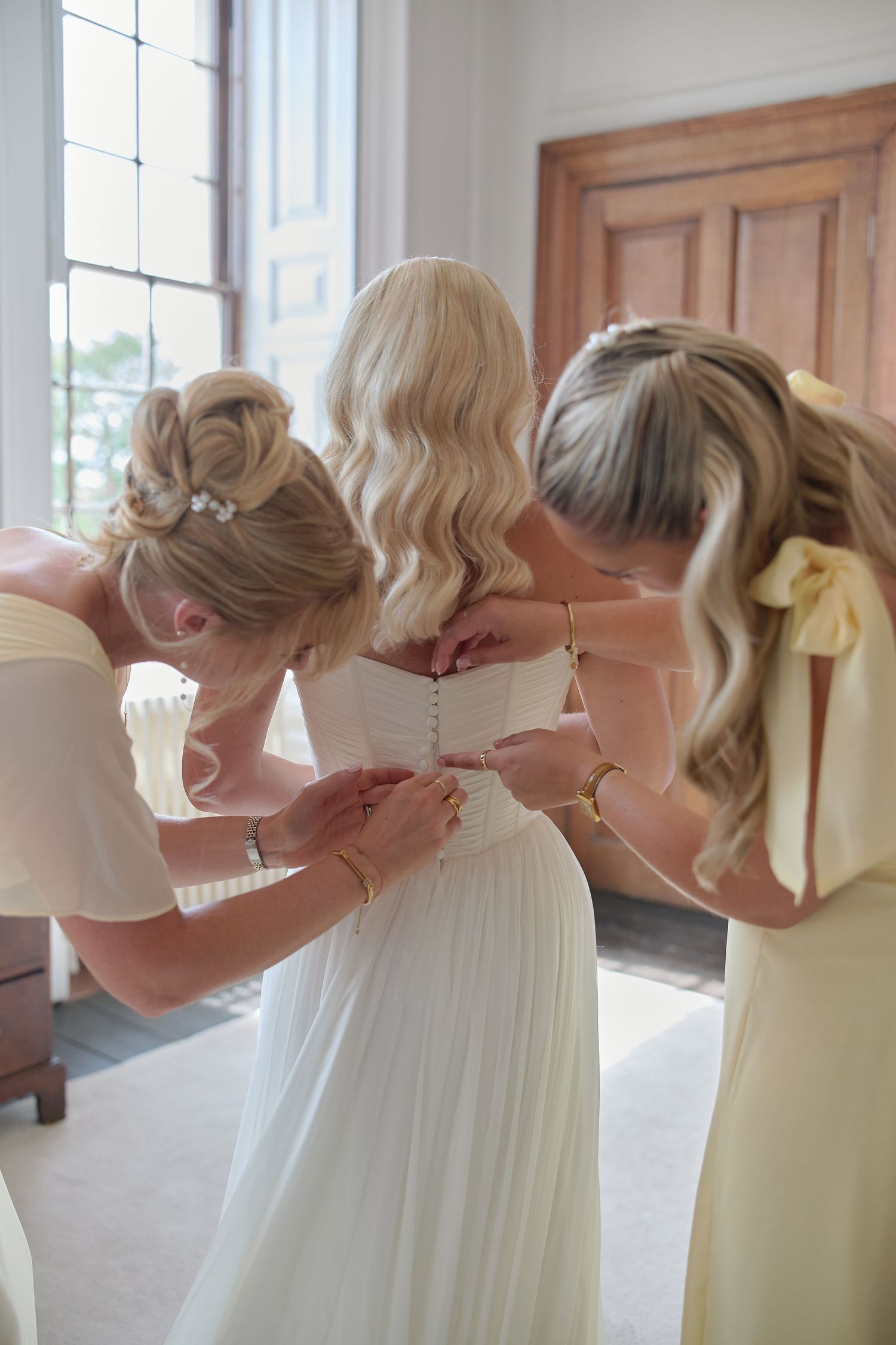 Two bridesmaids in yellow dresses help a bride with her white wedding gown buttons near a bright window.