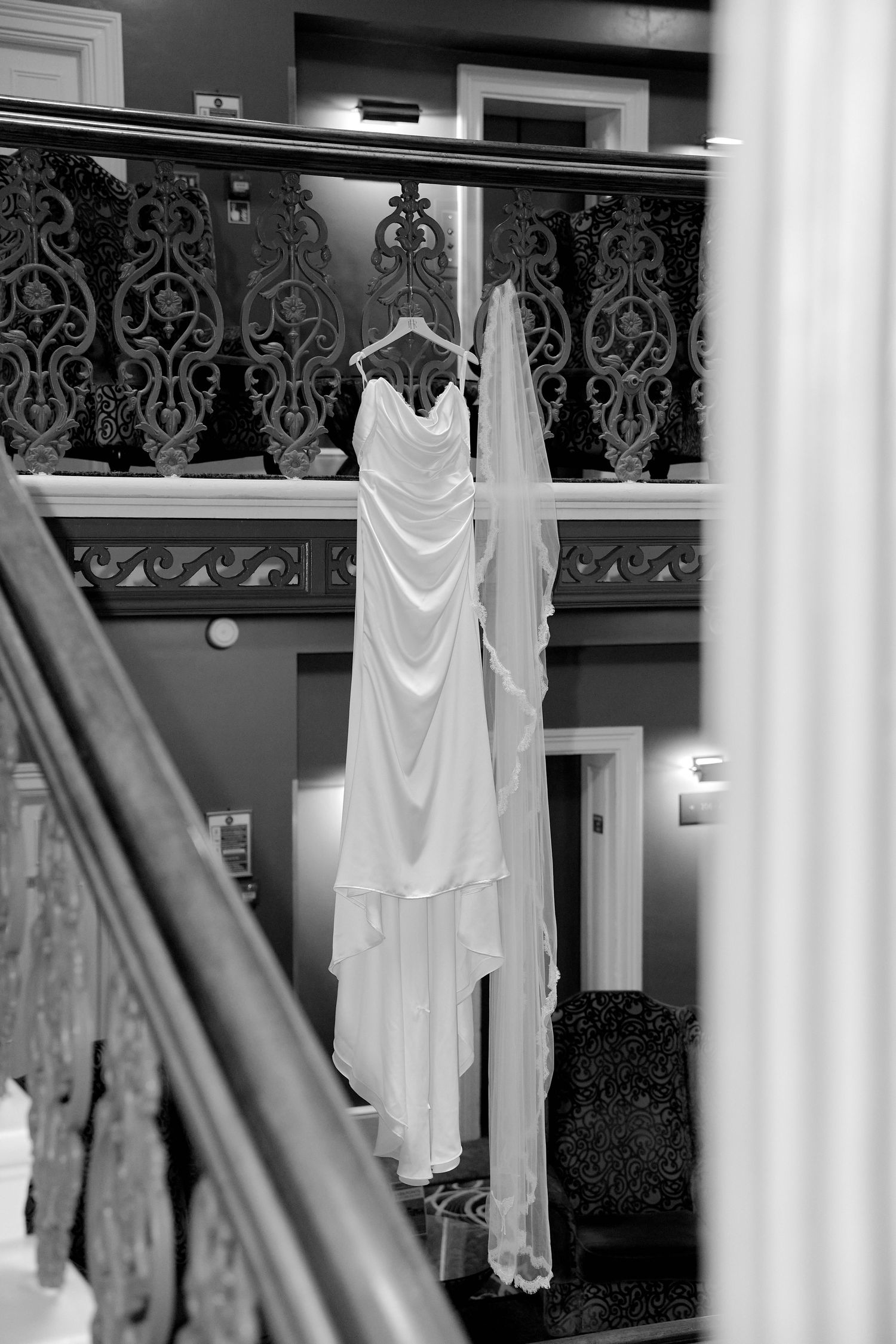 Black and white photo of an elegant wedding dress and veil hanging on a staircase railing in a grand venue.
