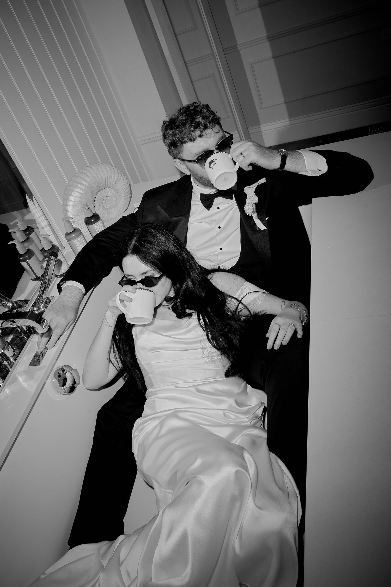 Black and white photo of bride and groom in formal wear drinking from cups, bride wearing sunglasses.