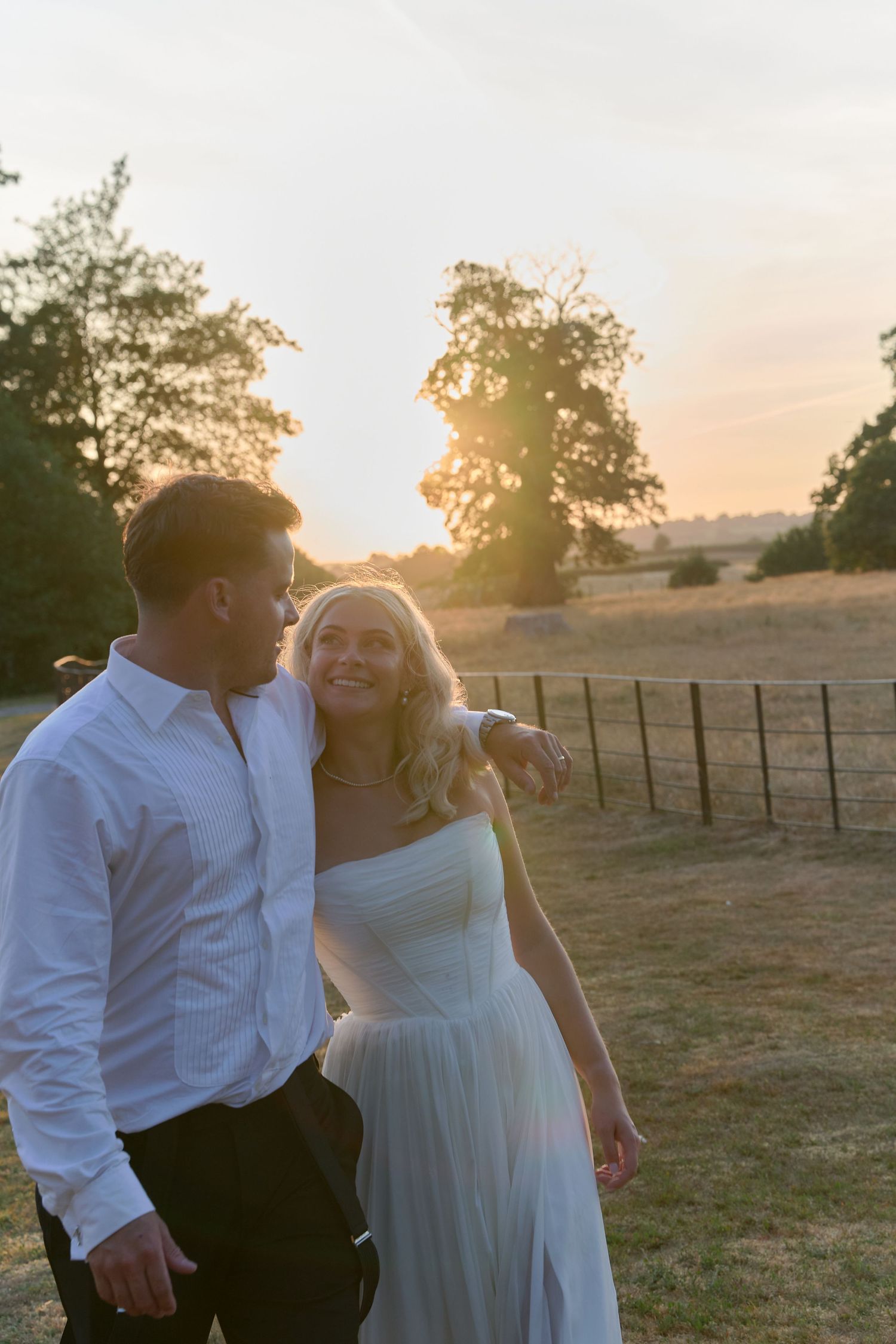 Couple walking at golden hour sunset, bride in white strapless gown, groom in white shirt, smiling together outdoors.