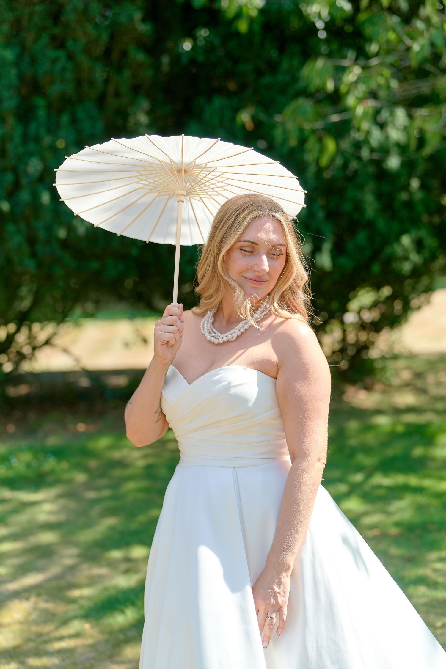 Blonde bride in strapless white gown holds ivory parasol outdoors, wearing pearl necklace in sunny garden setting.