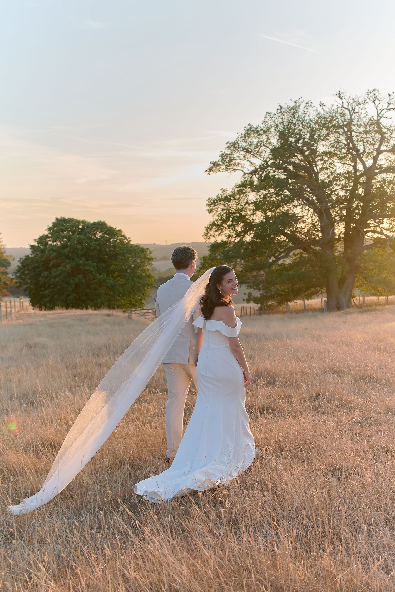 Bride and groom walk through golden field at sunset, her veil flowing in the breeze near a large oak tree.