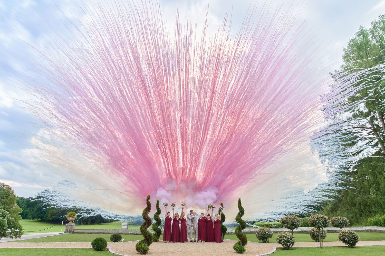 Bridal party in burgundy gowns beneath a stunning pink fireworks explosion at an elegant outdoor garden wedding.