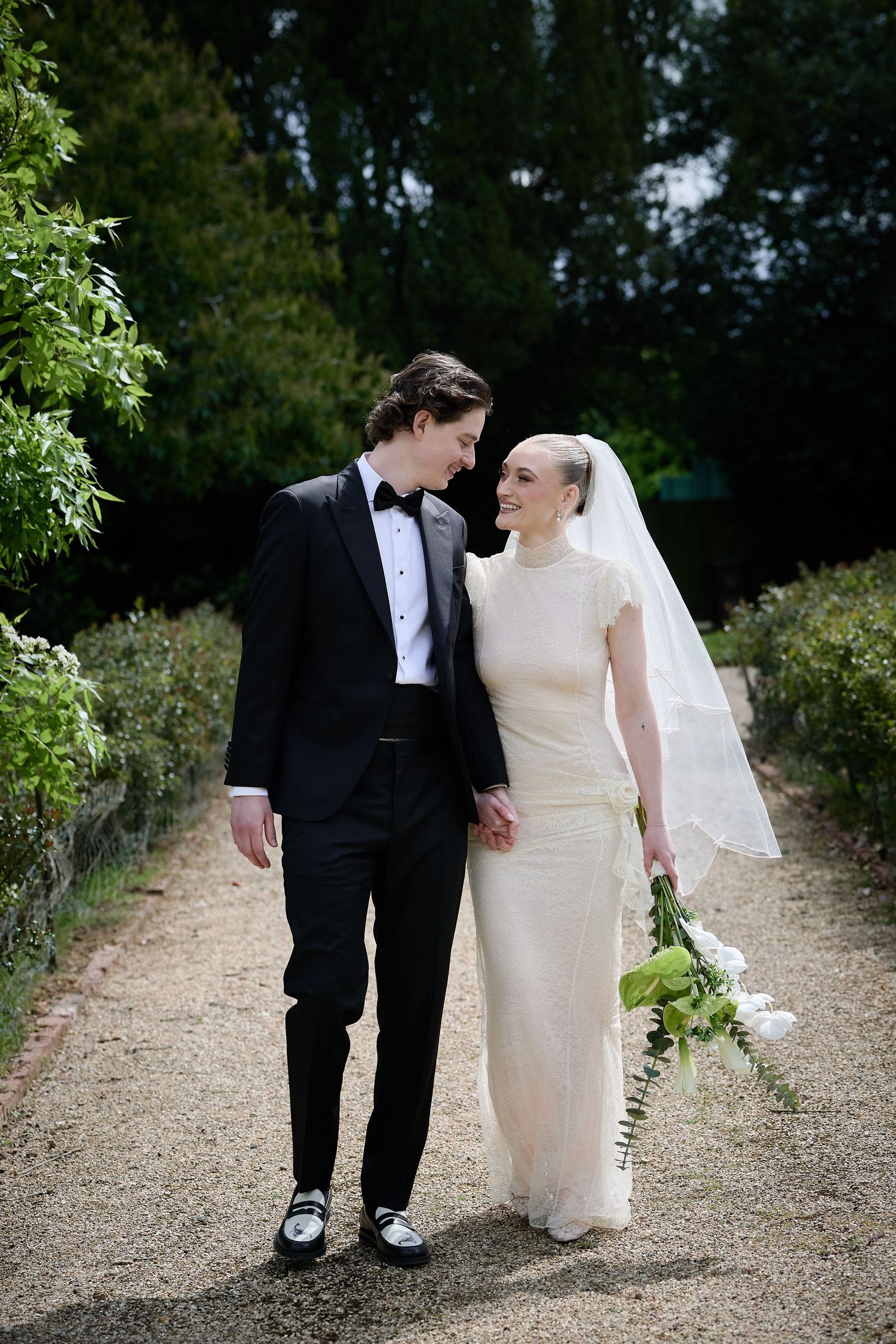 A bride and groom walk hand in hand along a garden path, smiling at each other on their wedding day.