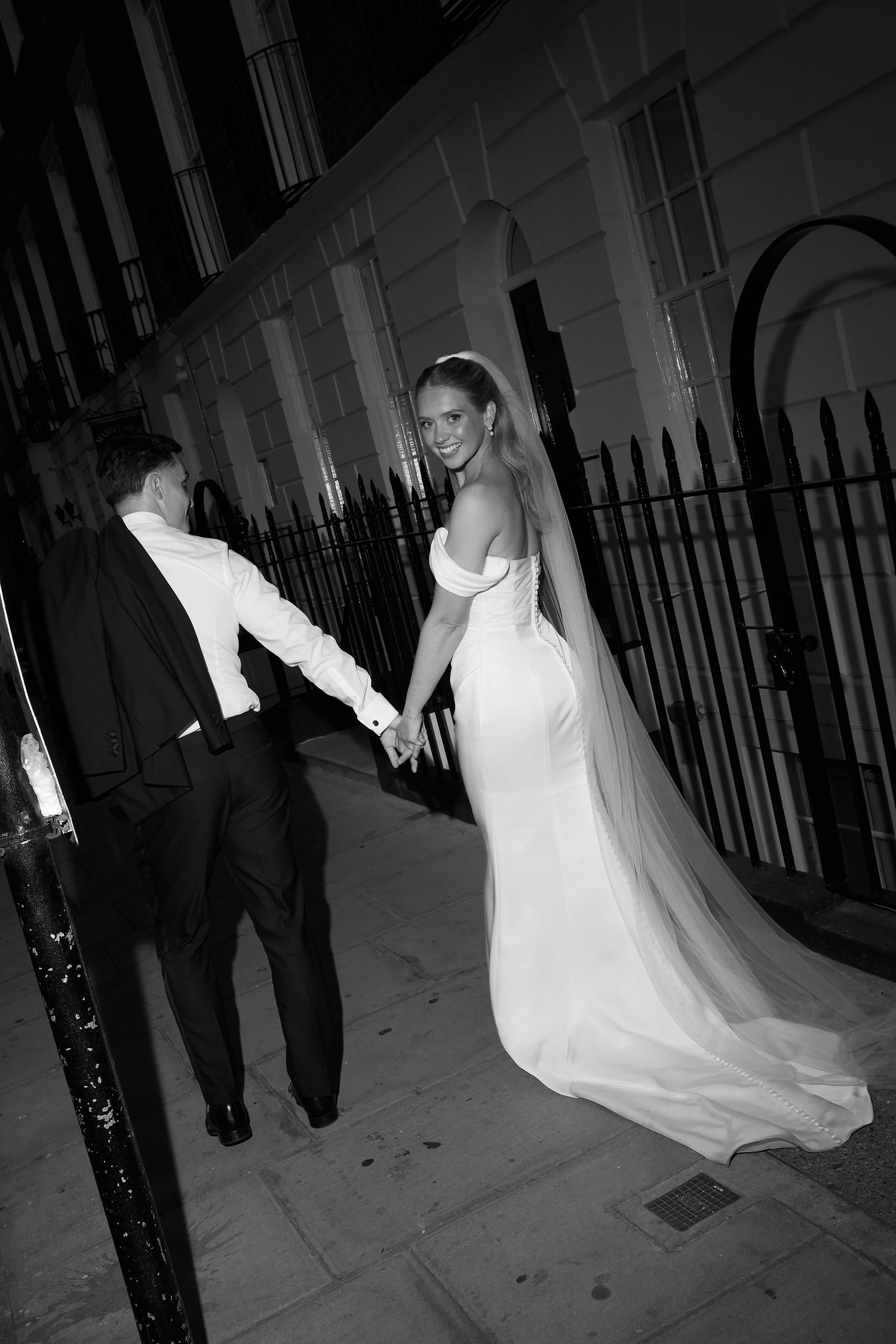 Black and white photo of bride in off-shoulder gown with veil holding hands with groom in tuxedo on city street at night.