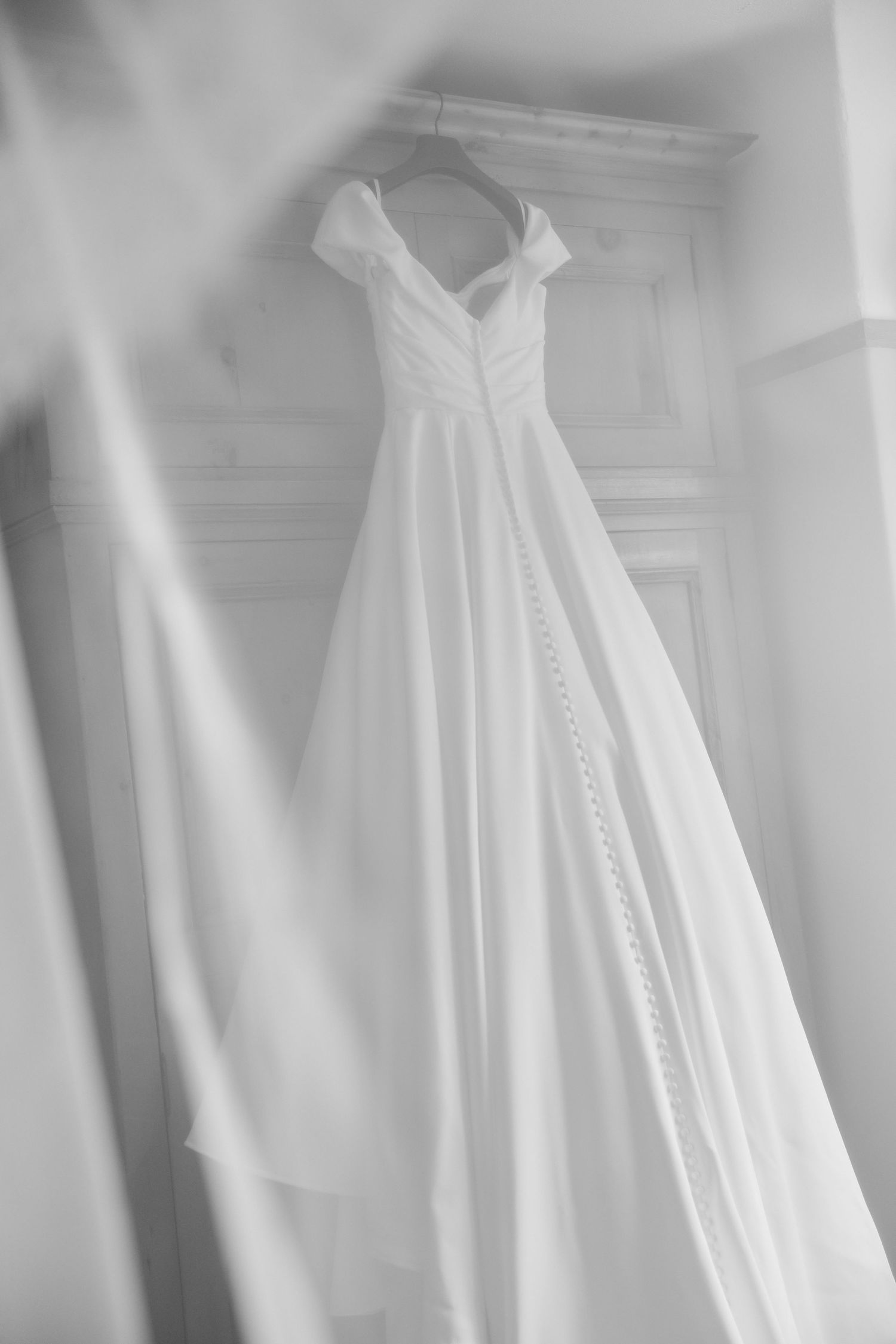 Elegant white wedding dress with off-shoulder straps and flowing skirt hanging on a hanger in black and white.