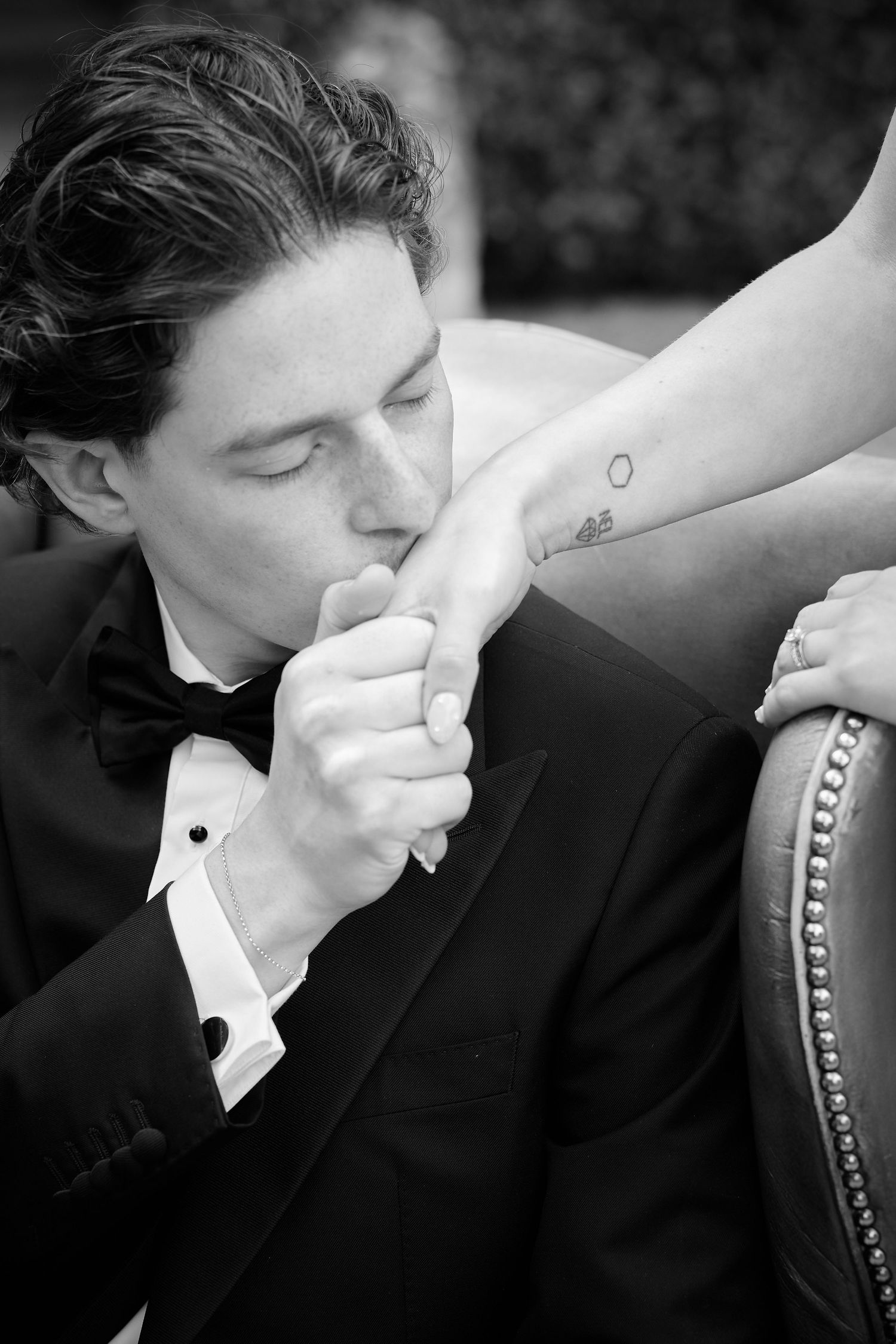Black and white photo of a groom in a tuxedo kissing his bride's hand at a wedding.