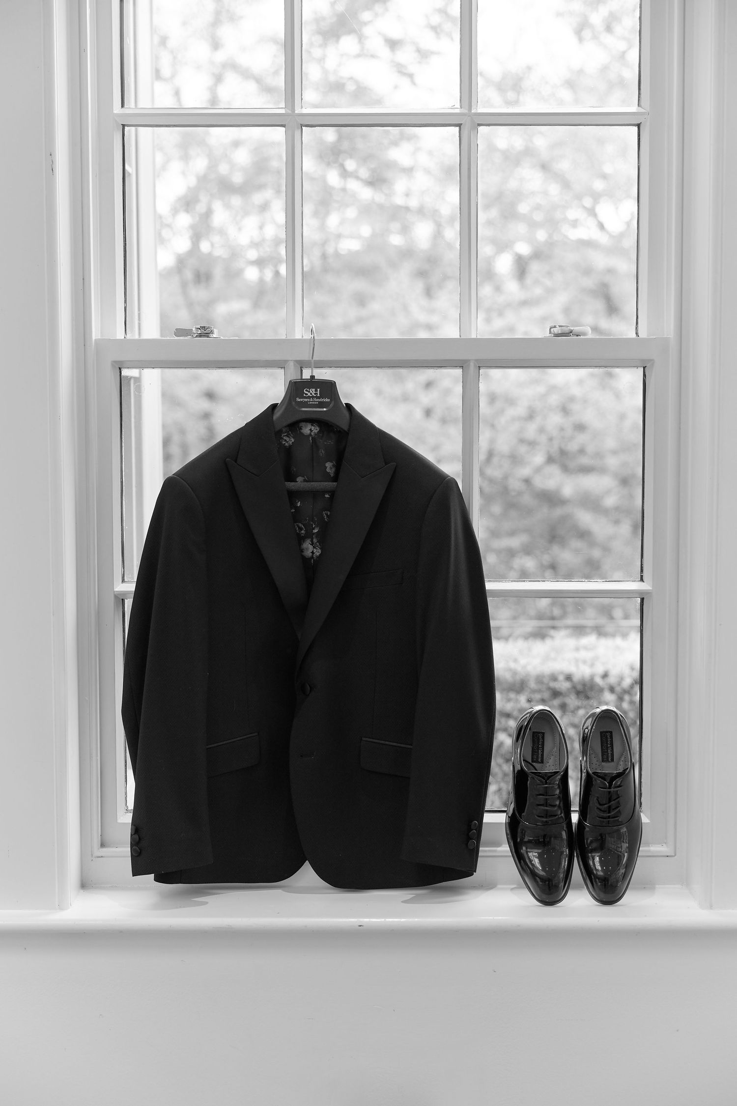 Black suit jacket on hanger and dress shoes displayed on window sill in elegant black and white photo.