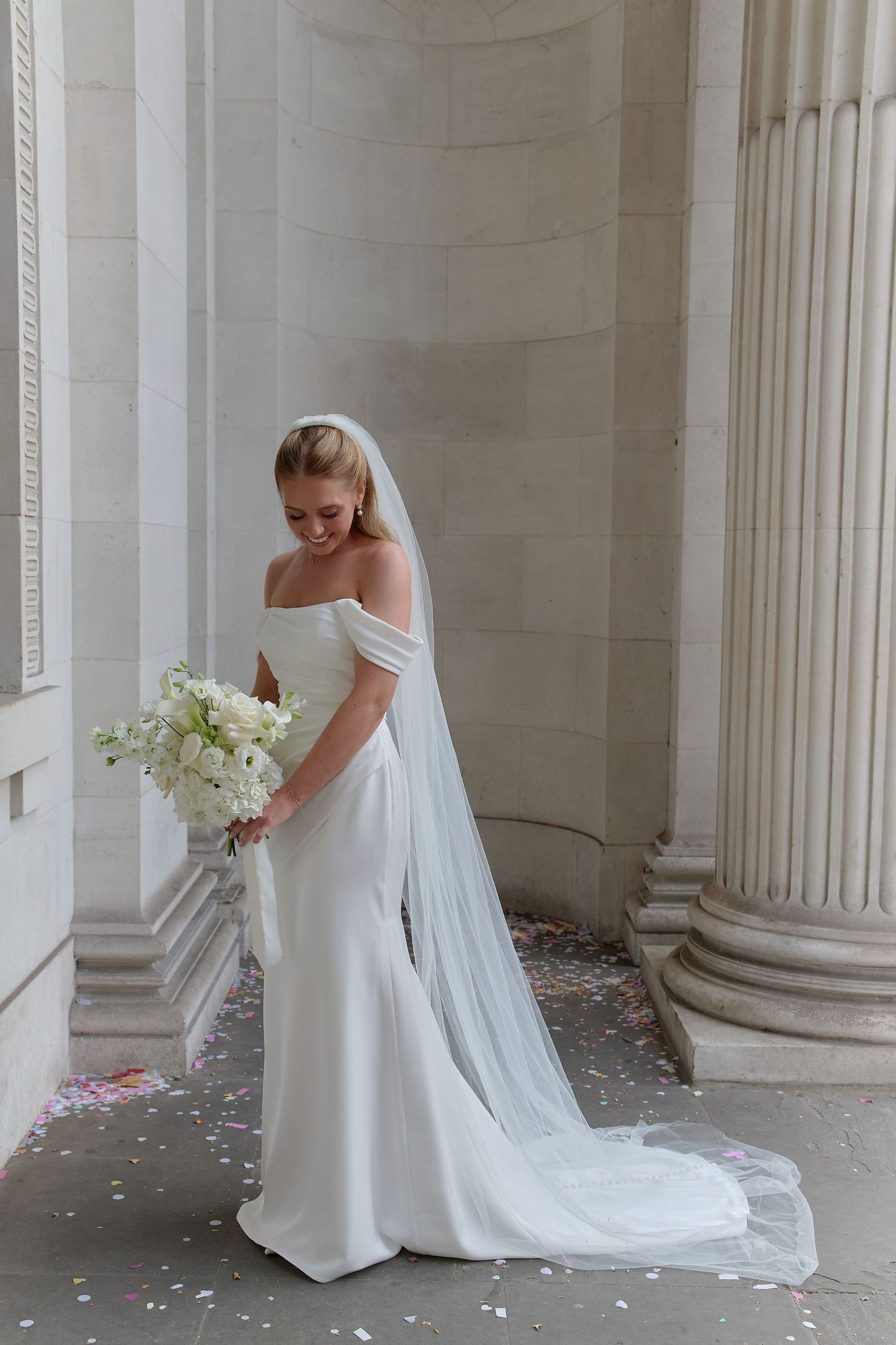 Bride in off-shoulder white gown with long veil holds white bouquet among stone columns with confetti.