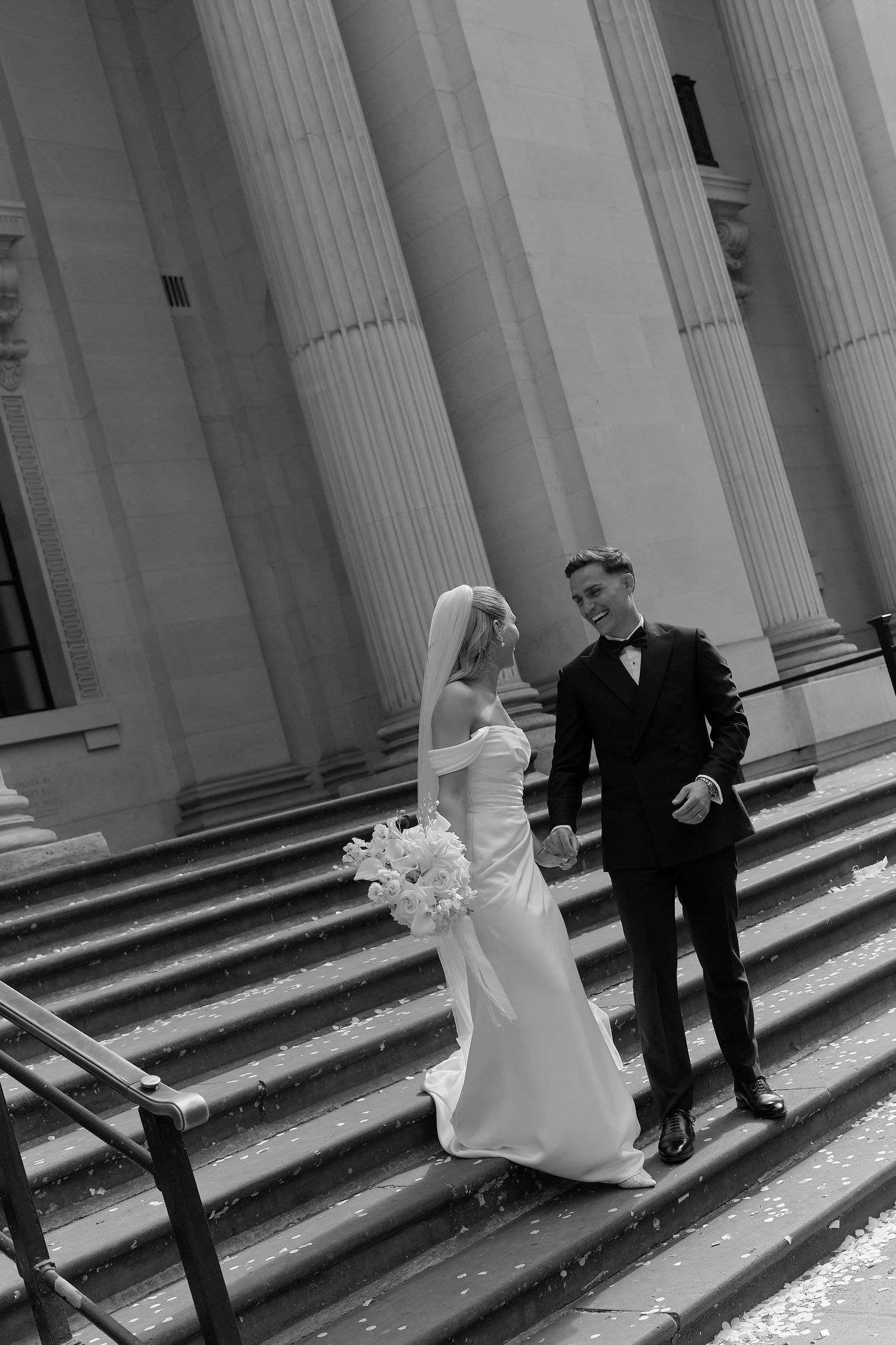 Black and white photo of a bride and groom holding hands on church steps with grand columns behind them.