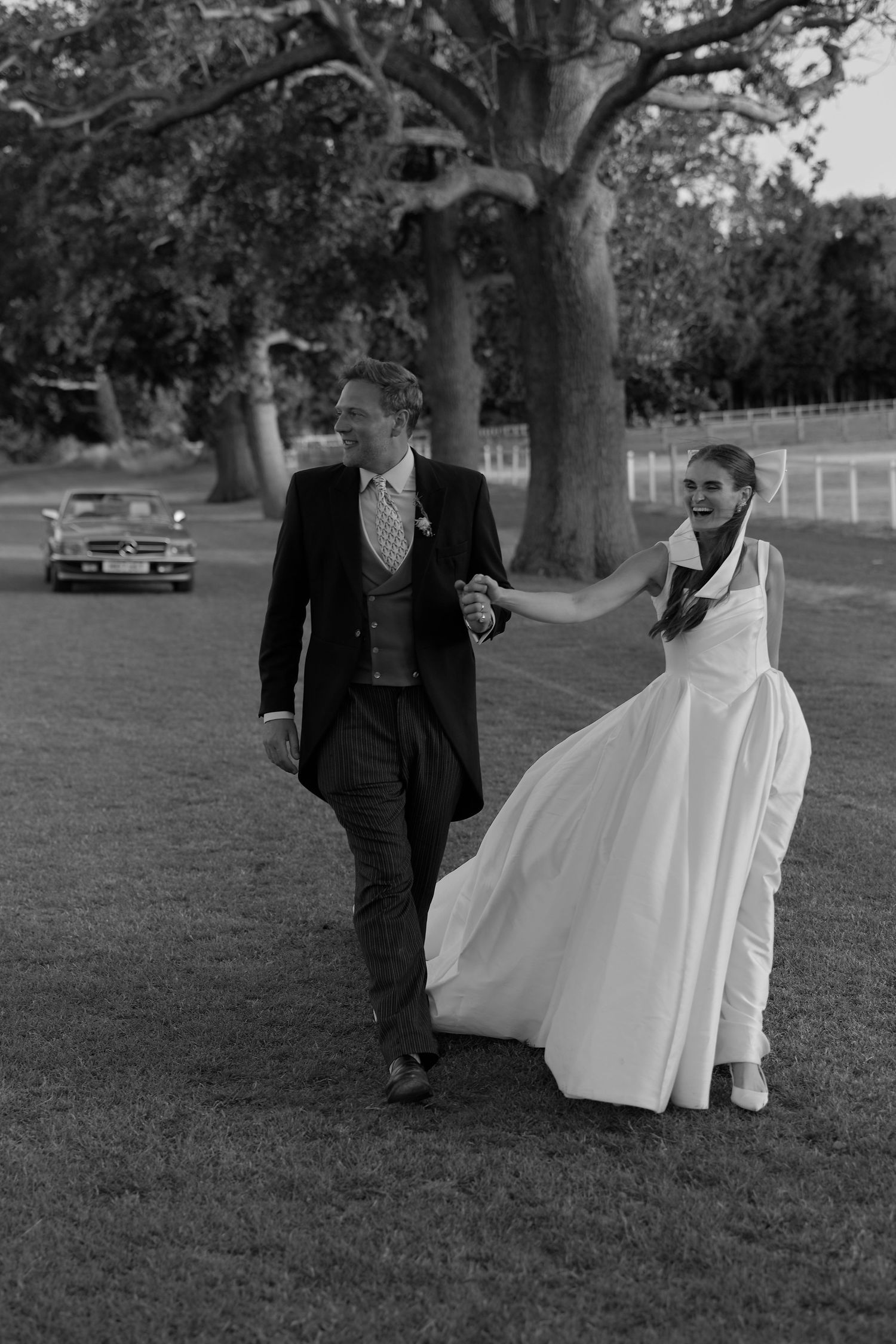 Black and white photo of a bride and groom walking hand in hand on grass, with a vintage car and trees in the background.