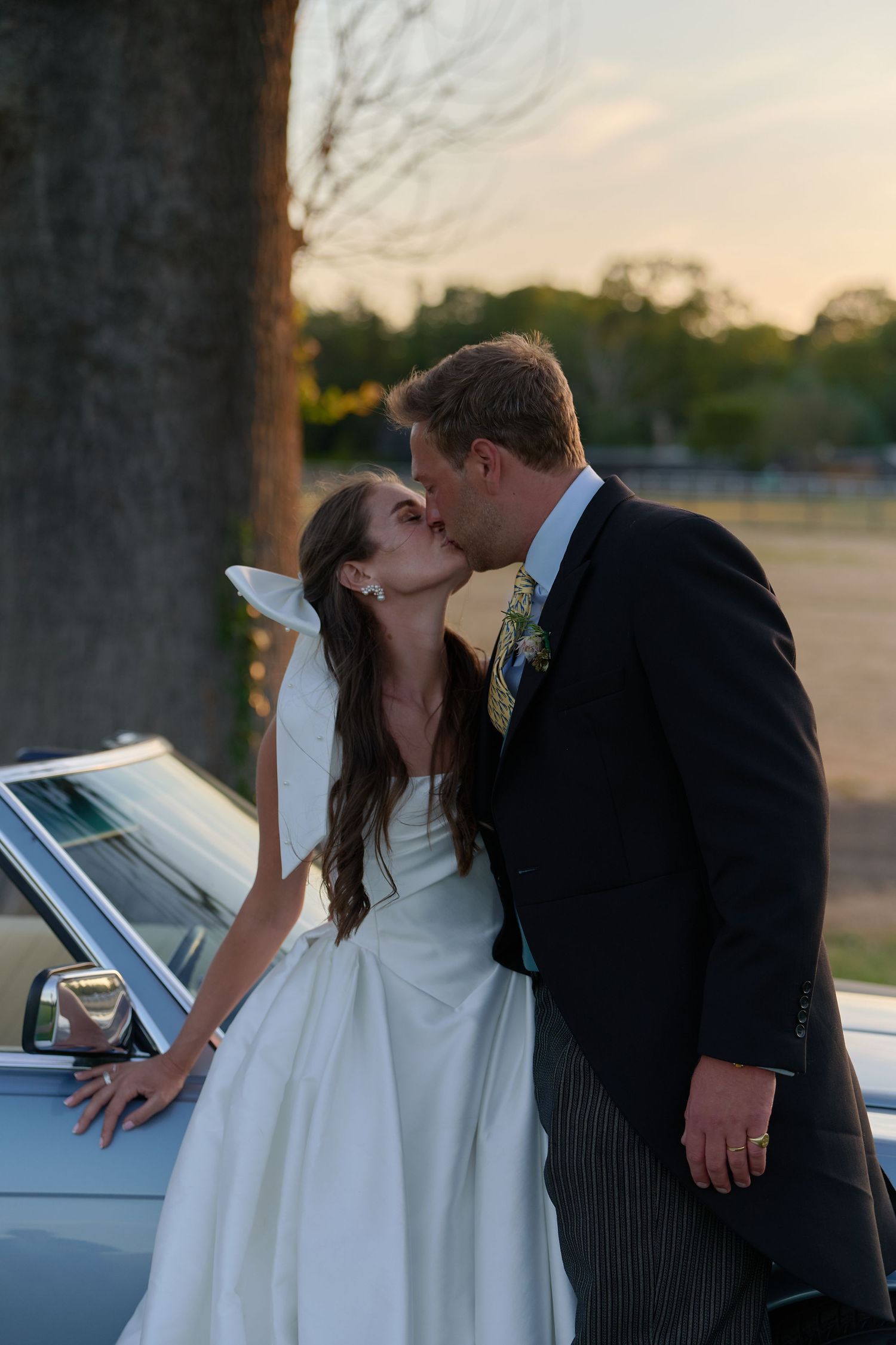 Bride and groom sharing a kiss beside a vintage convertible car at golden hour.
