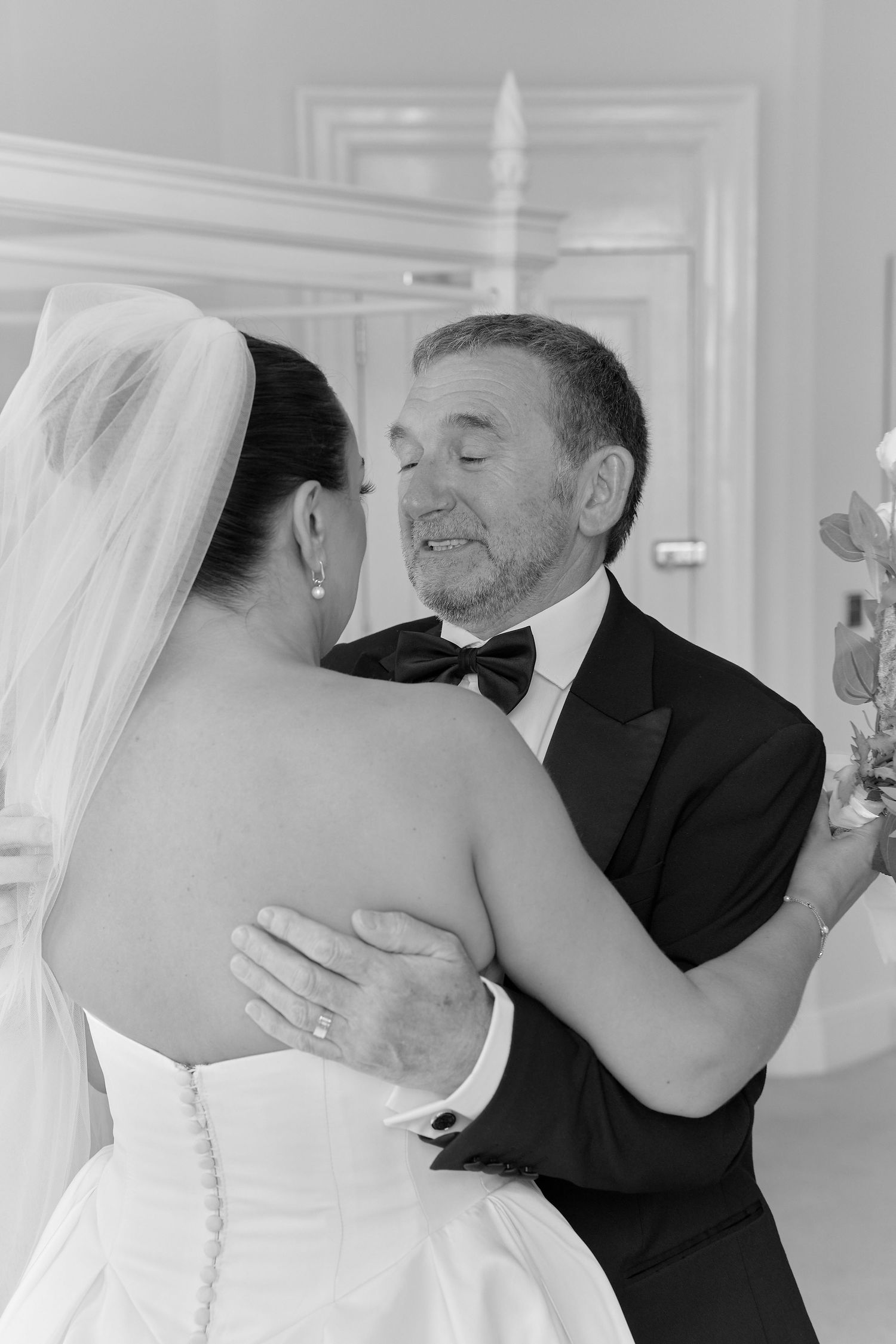 Black and white photo of a bride and older man in tuxedo sharing an emotional dance embrace at a wedding.
