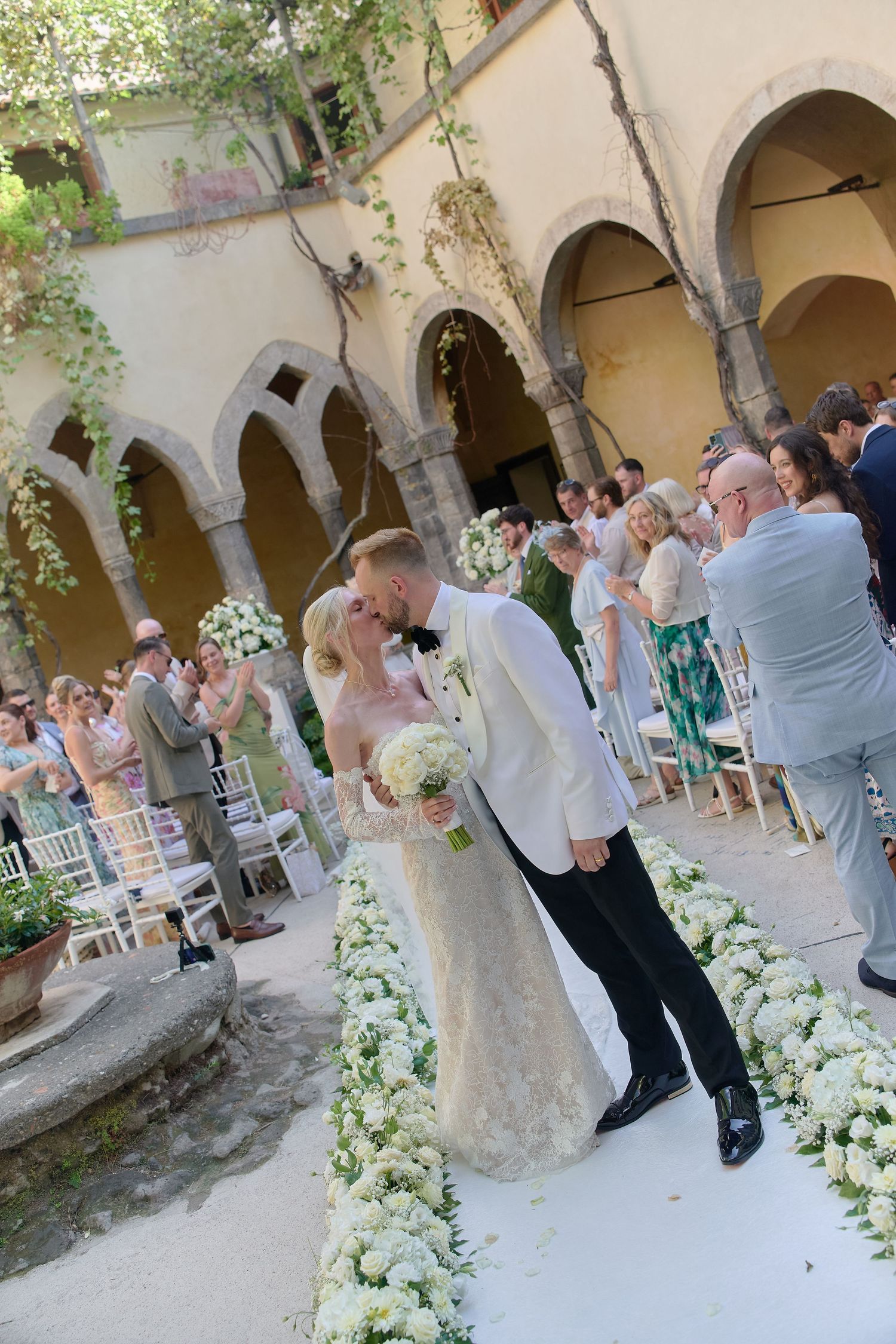 Bride and groom share a kiss while walking down a floral aisle at an outdoor wedding in a historic Italian courtyard.