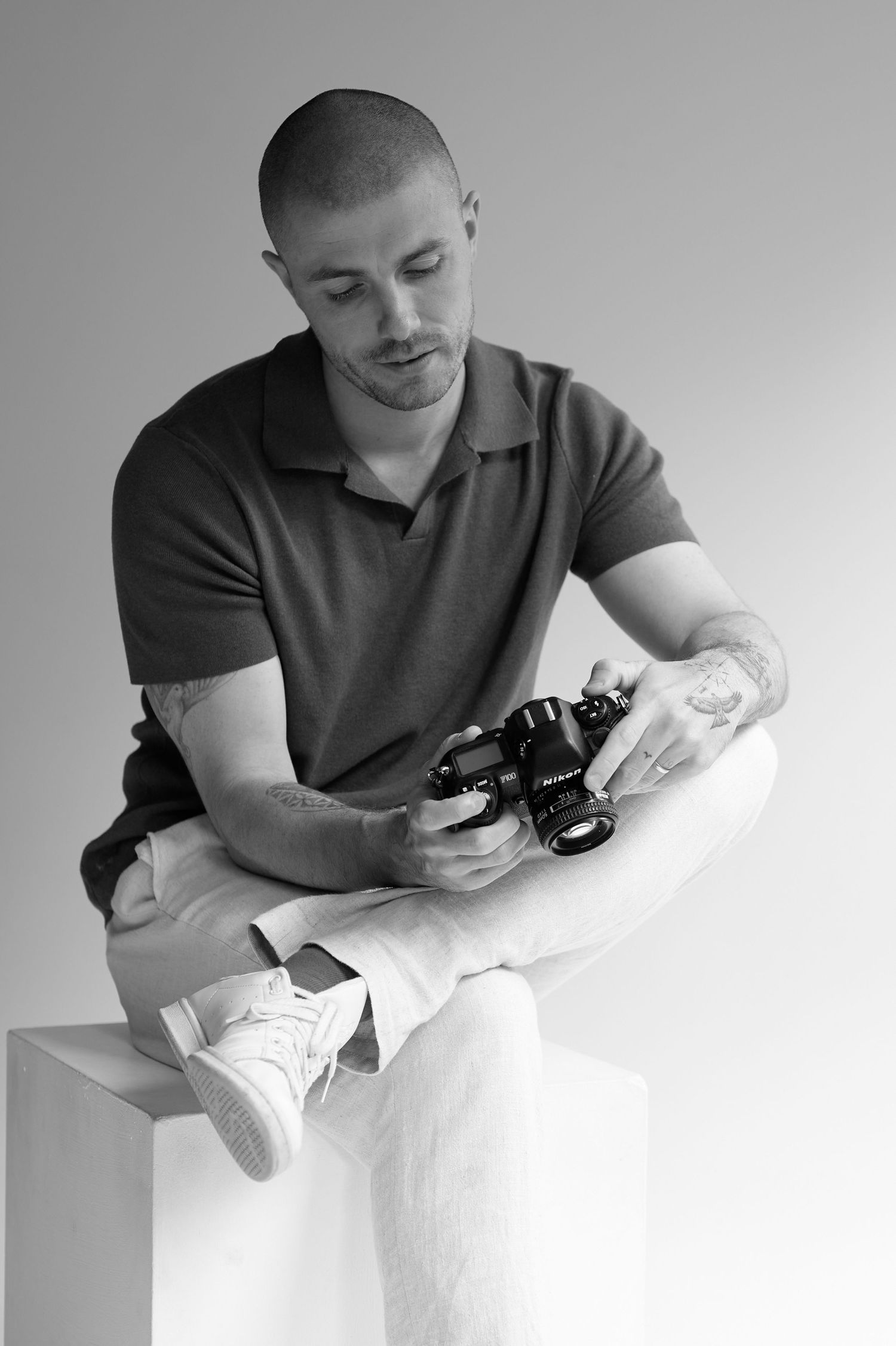 Photographer examining camera while seated on white cube in black and white studio photo.