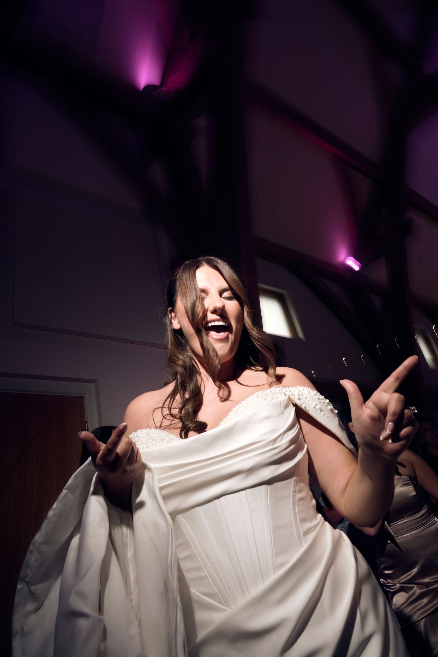 Bride in white off-shoulder gown laughing and dancing joyfully at wedding reception with purple uplighting.