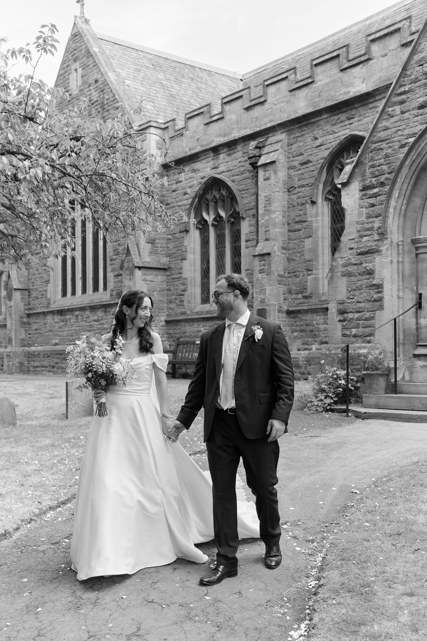Black and white photo of a bride and groom holding hands outside a historic stone church on their wedding day.