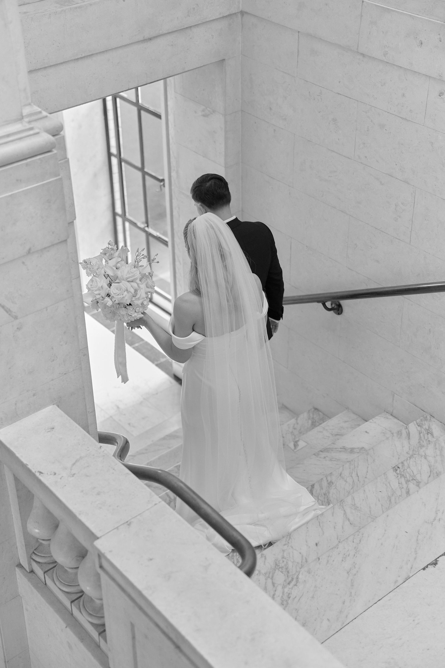 Aerial black and white photo of bride and groom ascending marble staircase, bride holding bouquet.