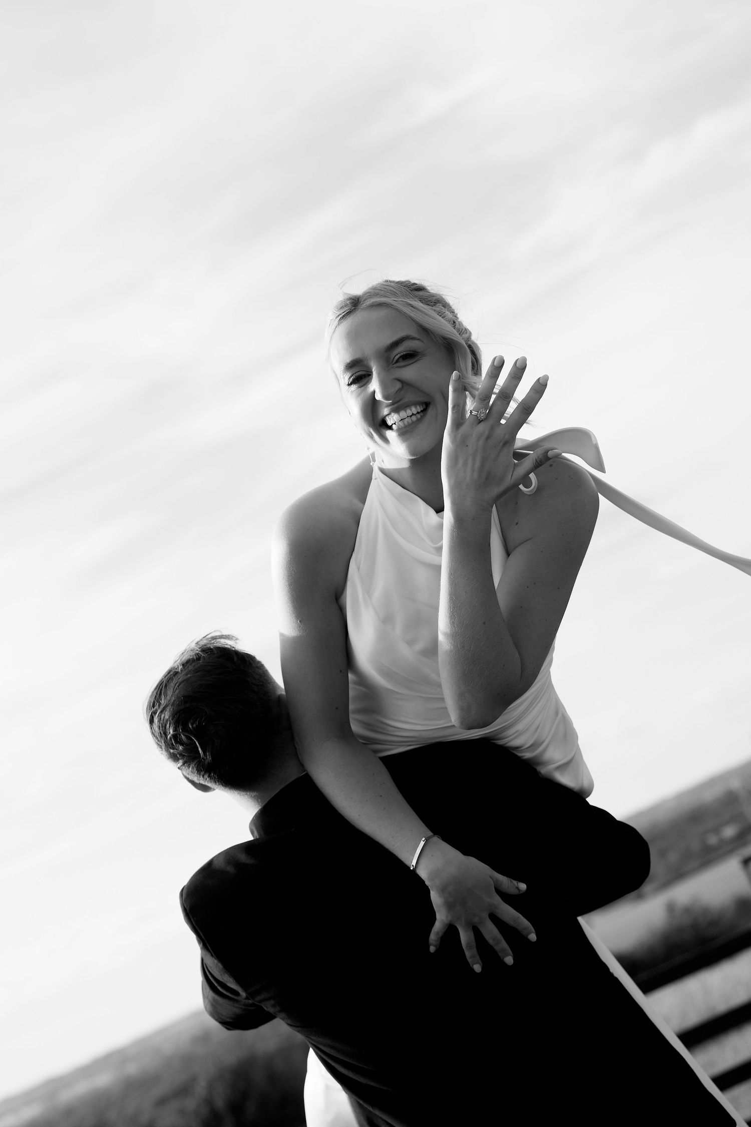 Black and white photo of a joyful woman being lifted by a man, showing off her engagement ring while laughing outdoors.