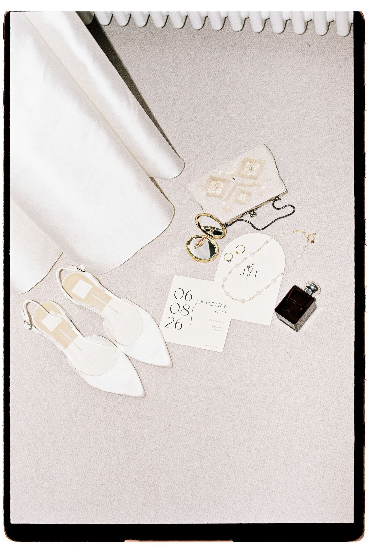 Elegant bridal flat lay featuring white slingback heels, clutch, gold jewelry, perfume bottle, and wedding invitation cards.