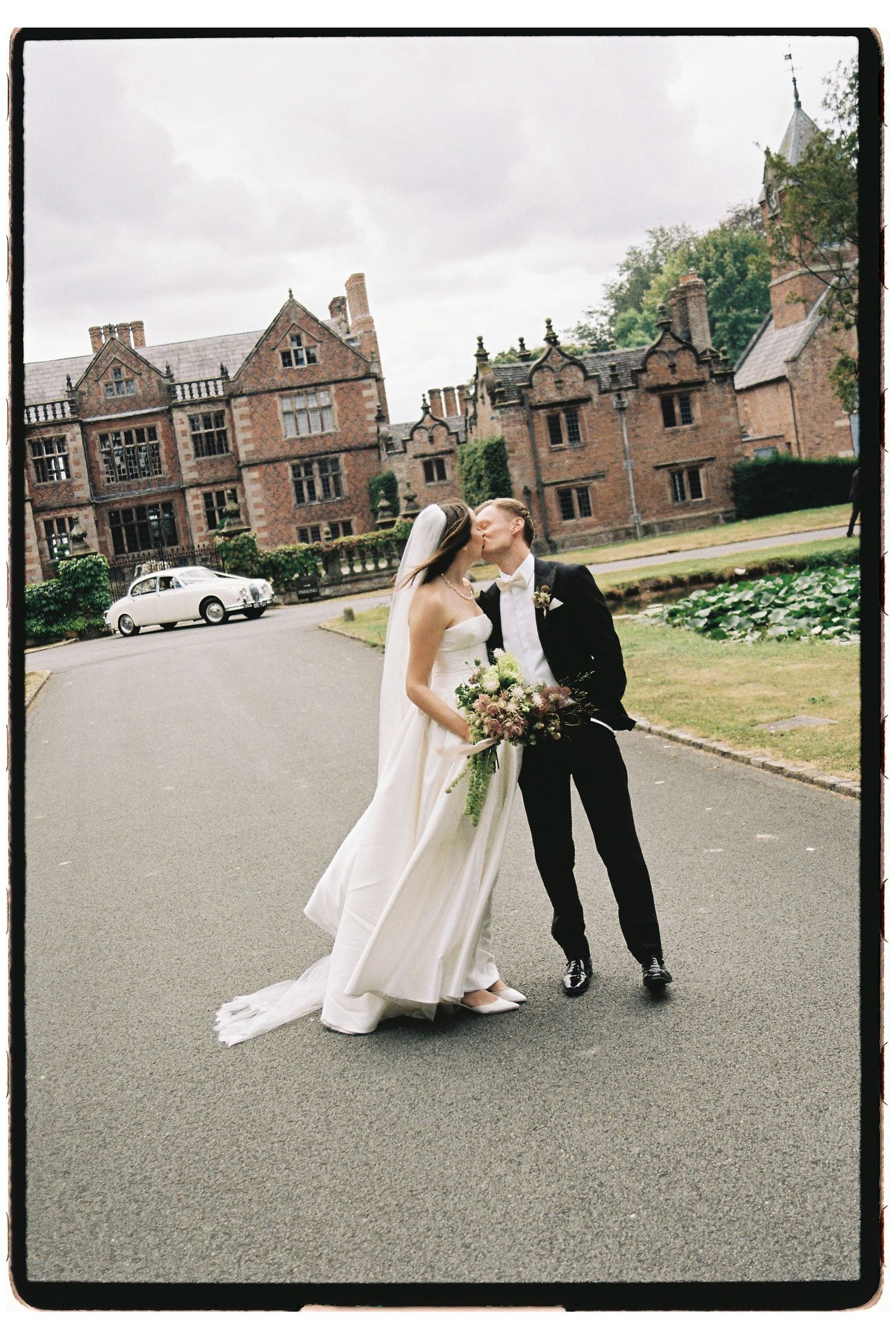 Bride and groom share a kiss outside a grand Tudor manor house on their wedding day, bride holding a bouquet.