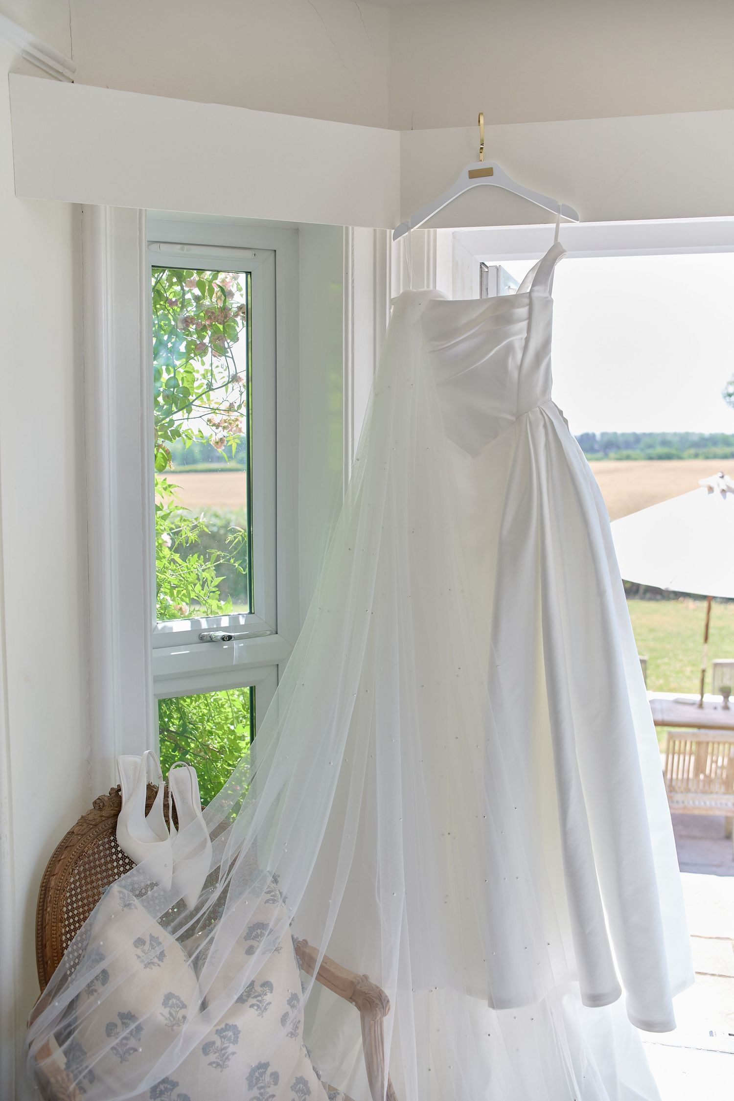 White wedding dress hanging by window with countryside view in bright natural light.