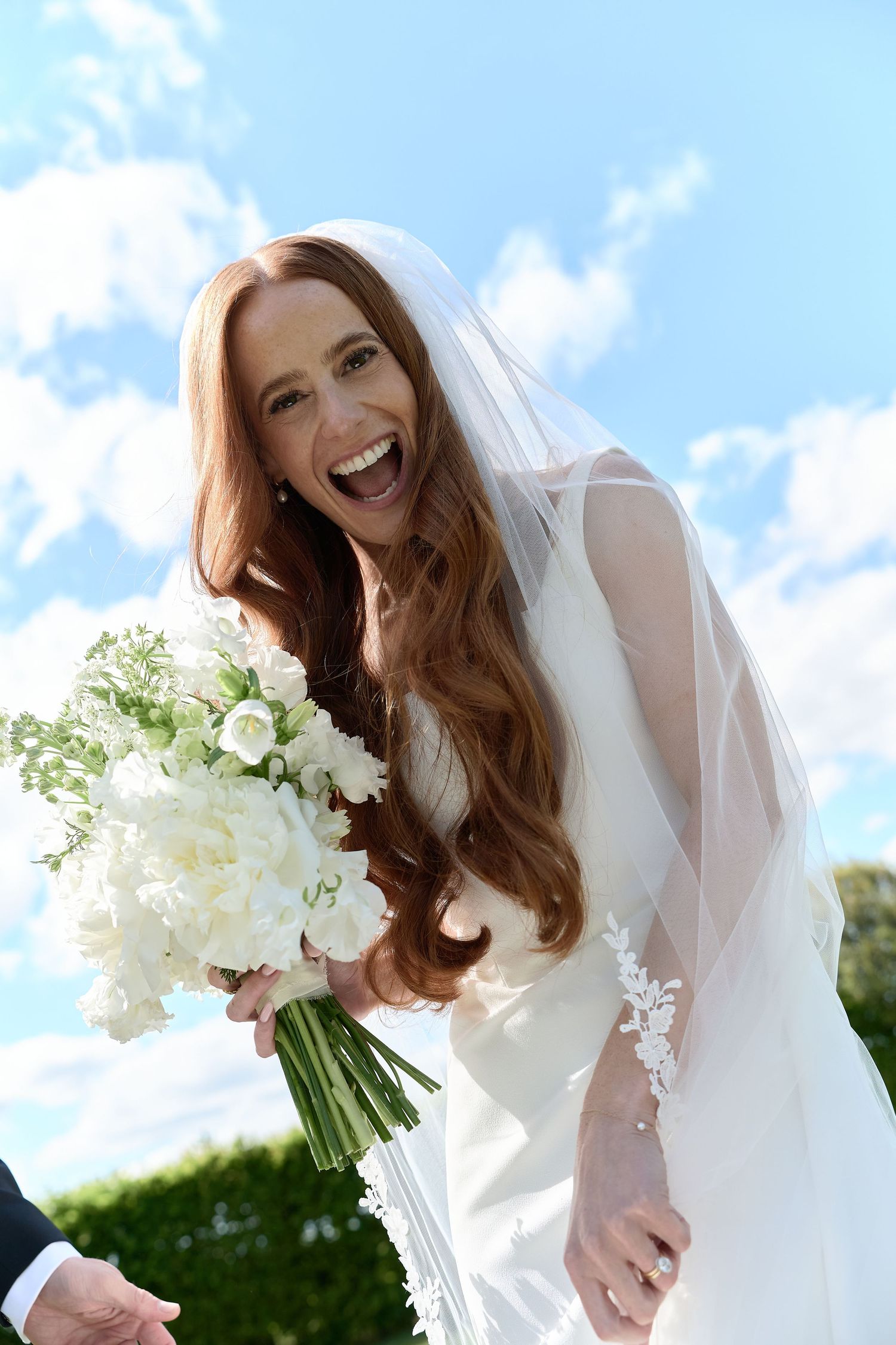 Joyful bride with long red hair laughing while holding a white floral bouquet on her wedding day.