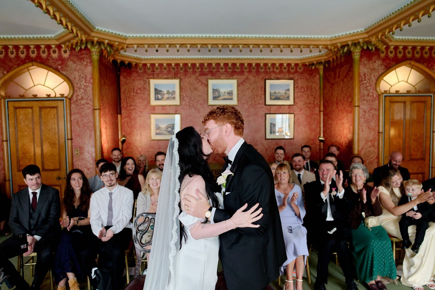 Bride and groom share first kiss at elegant indoor wedding ceremony with seated guests applauding in ornate red-walled venue.