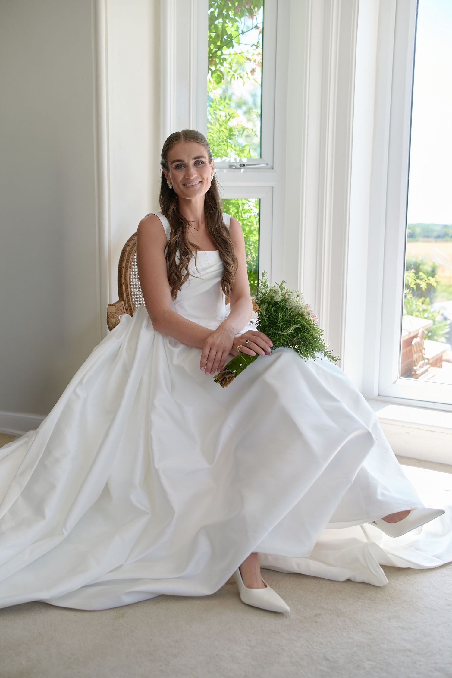 Bride in a white ball gown sitting by a window holding a green bouquet.