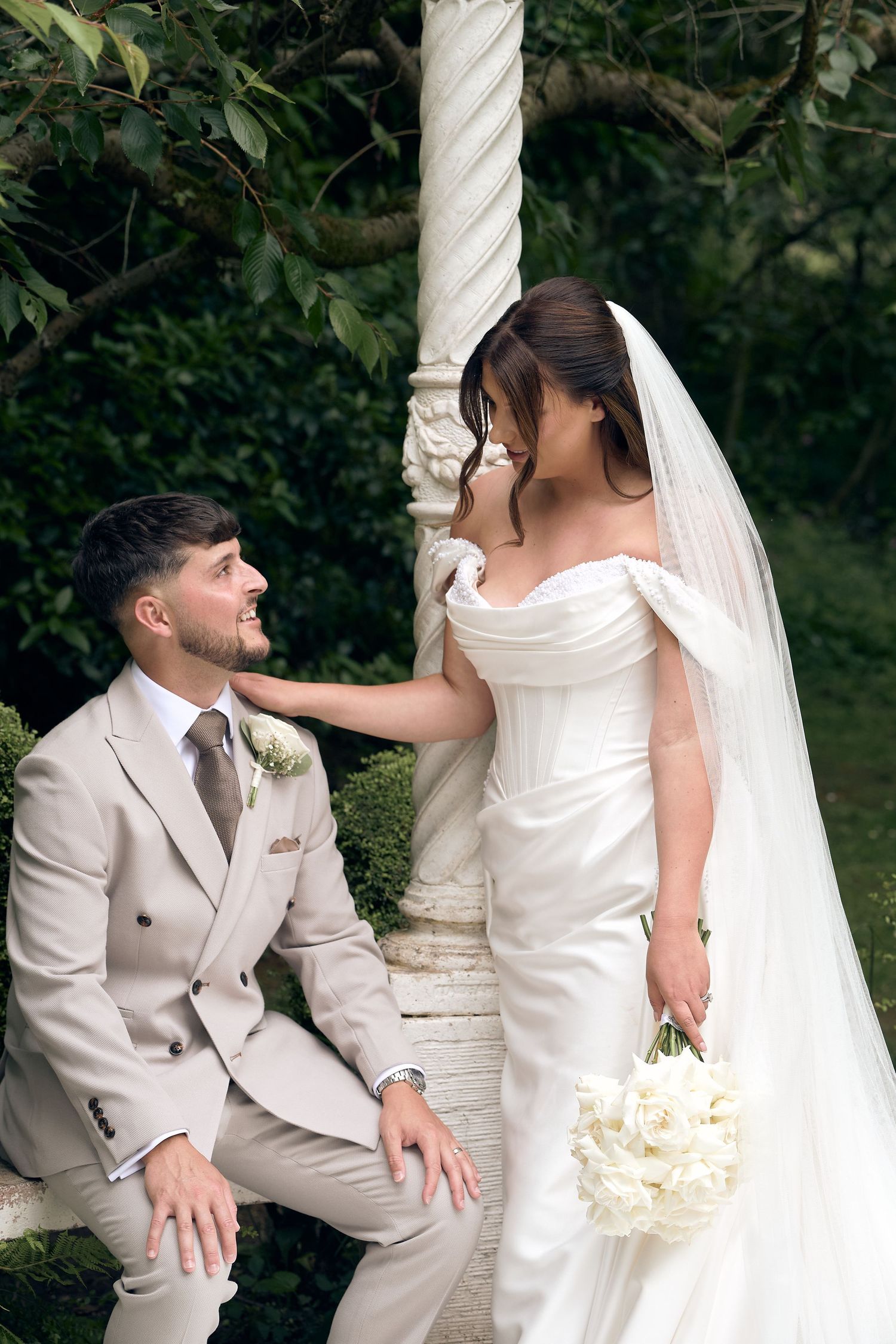 A groom in a beige suit sits smiling up at his bride in a white off-shoulder gown holding a bouquet in a garden.