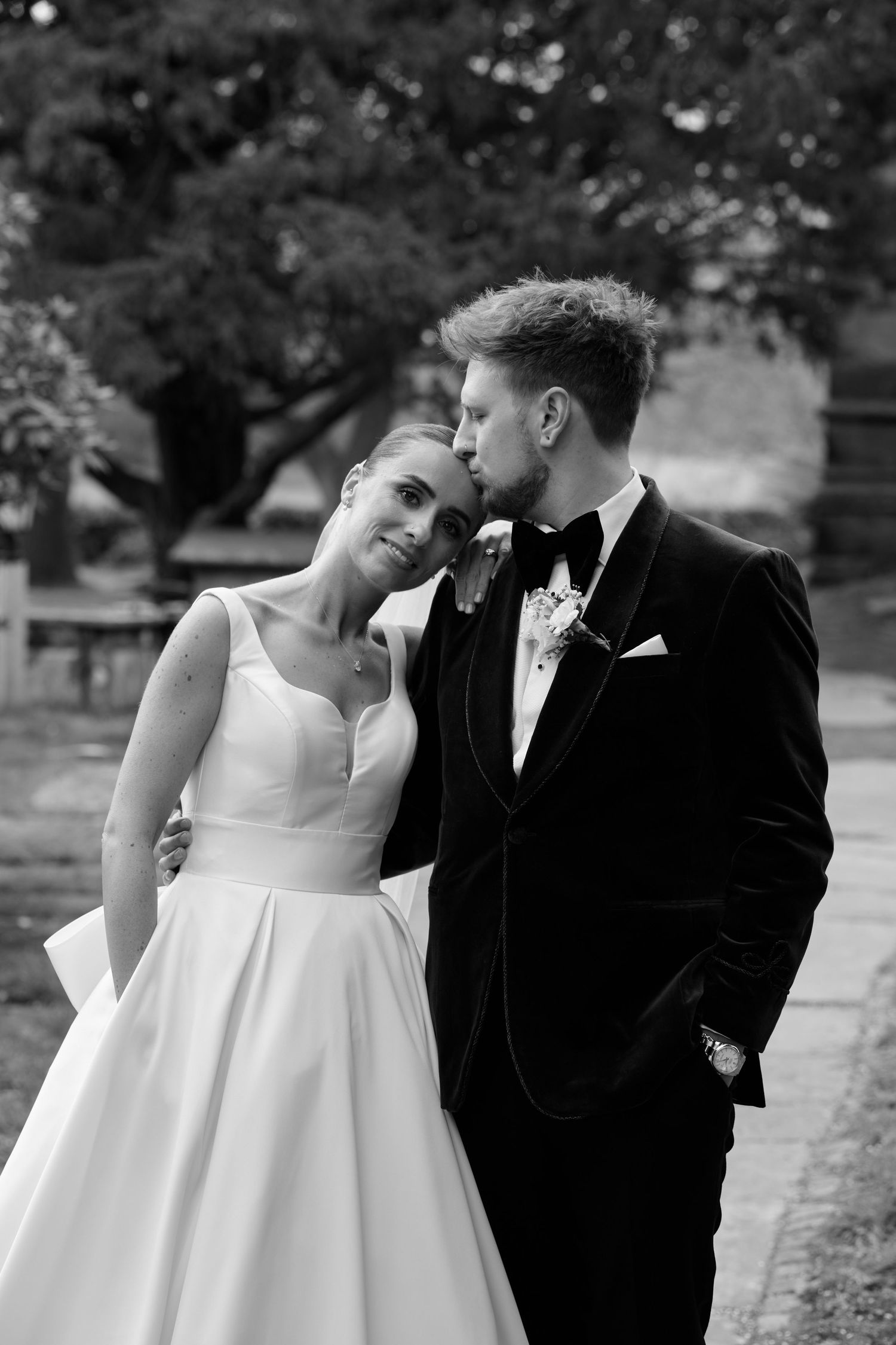 Bride and groom share tender moment outdoors, groom in velvet tuxedo kisses bride's temple.
