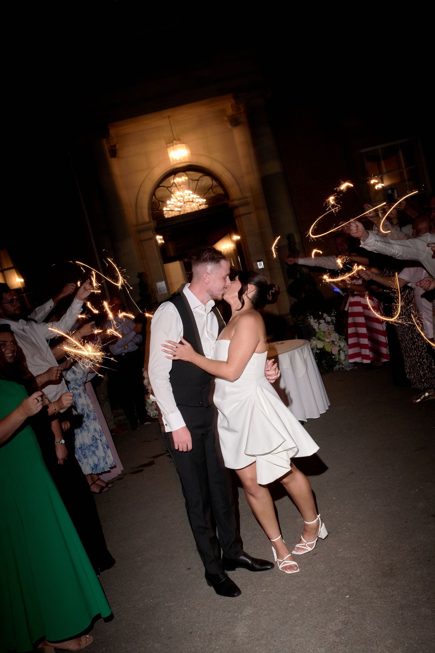 Newlyweds share a kiss during a sparkler send-off exit at night outside an elegant wedding venue.