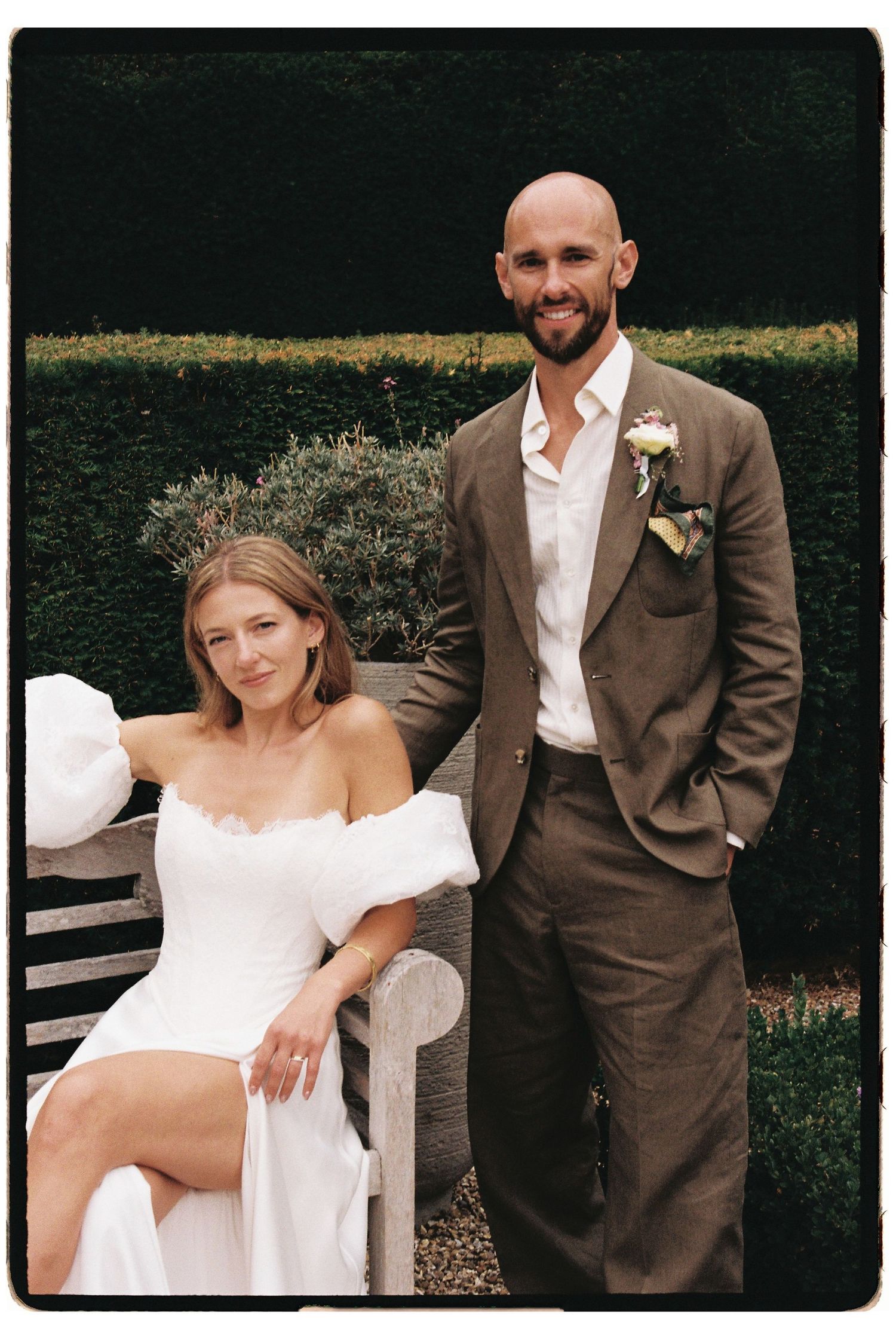 Bride in off-shoulder white dress seated on bench with groom in brown suit wearing boutonniere in garden setting.