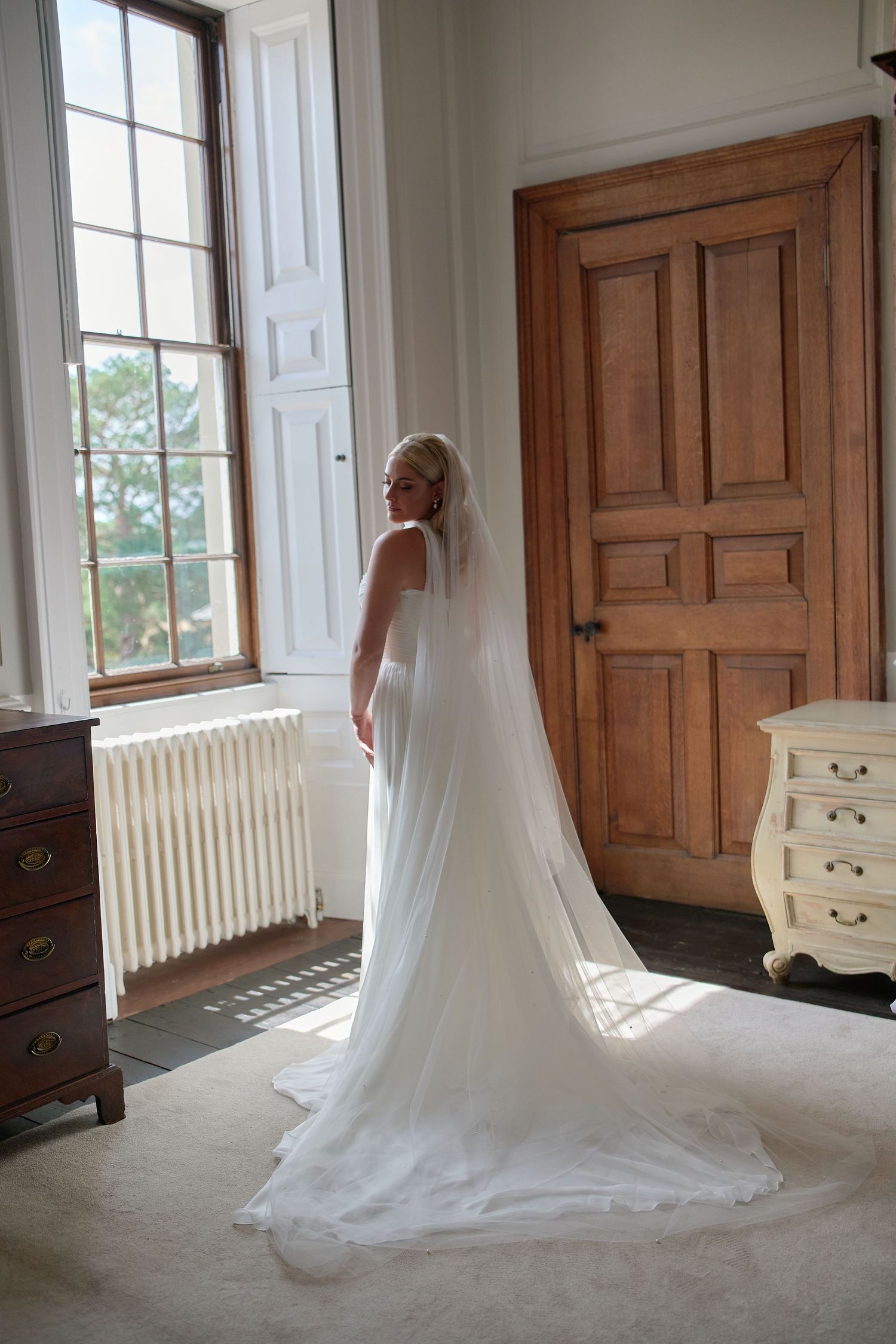 Elegant bride in a flowing white wedding gown with long veil standing by a sunlit window in a classic room.