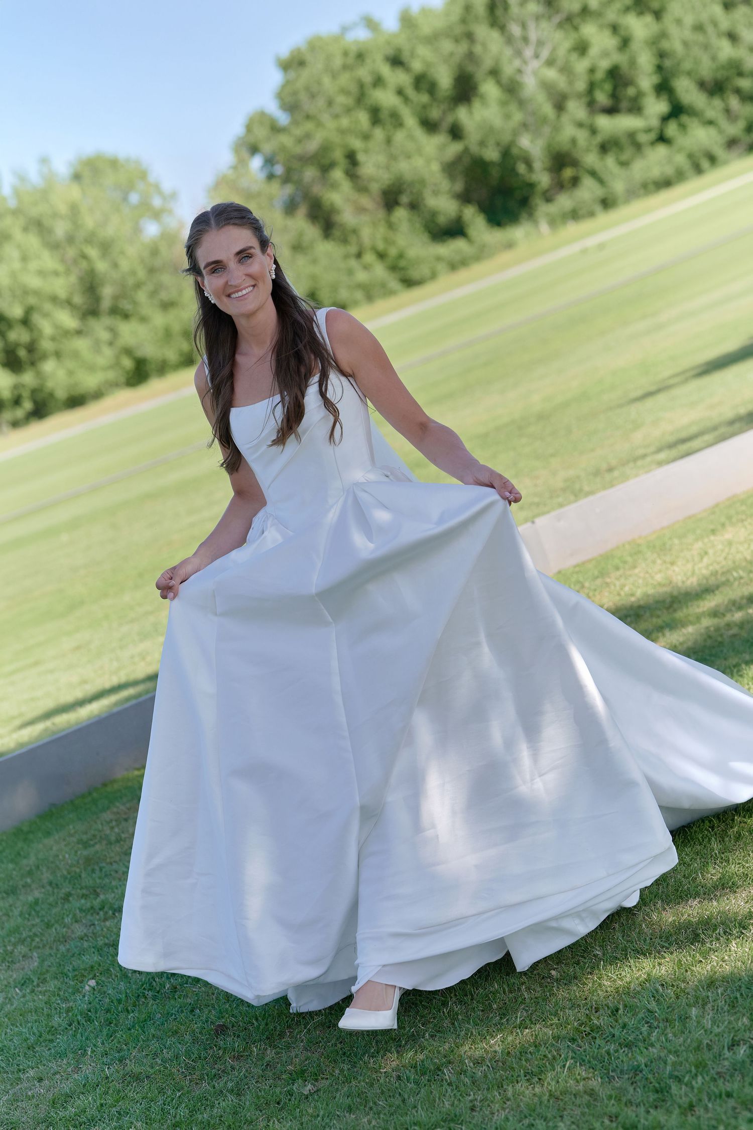 Smiling bride in a flowing white ball gown twirling on a green lawn outdoors.