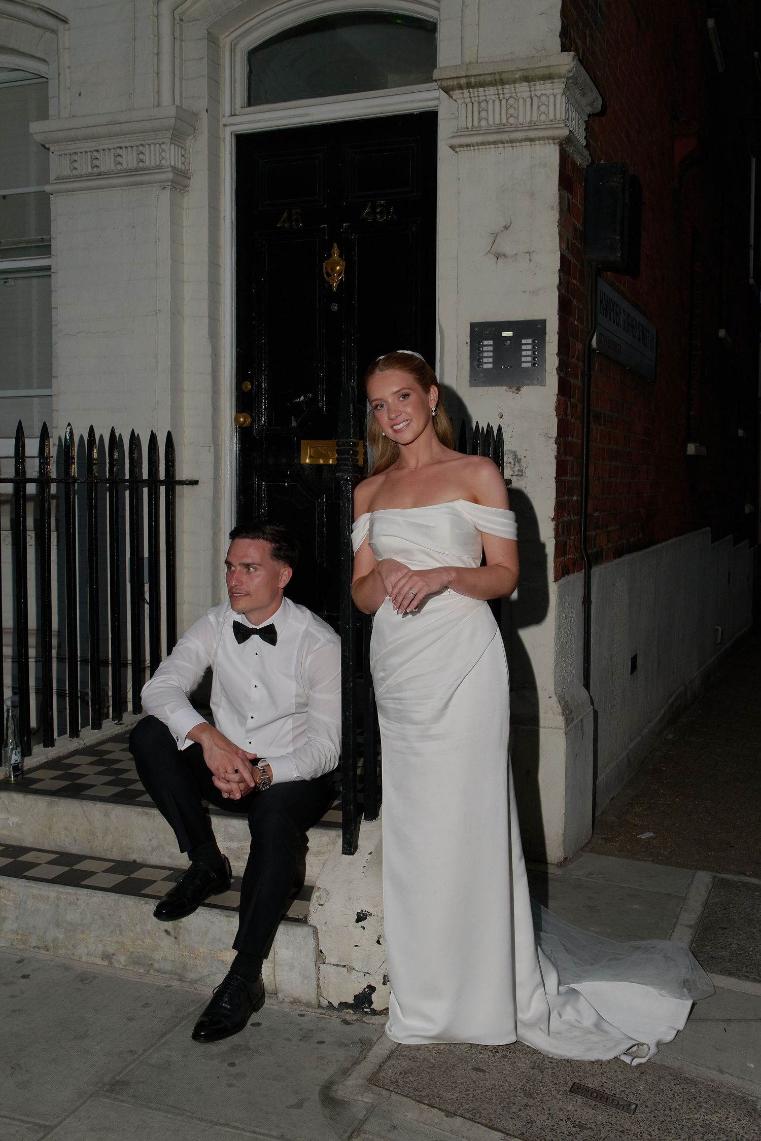 Bride in off-shoulder white gown stands beside groom in tuxedo on steps of elegant building at night.