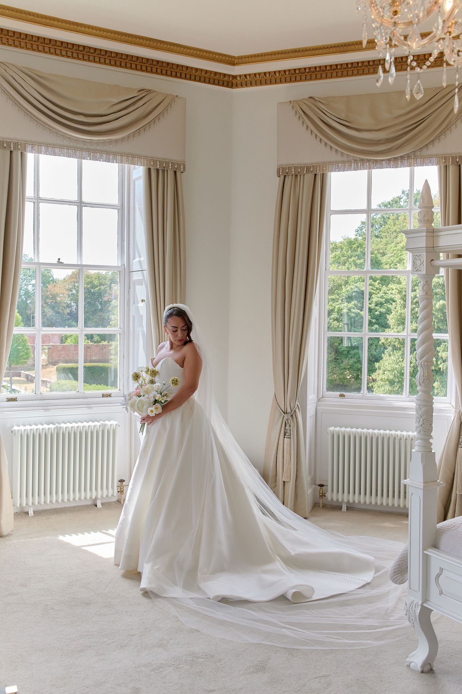 Bride in a white ball gown with long train holds bouquet in elegant room with tall windows and chandelier.