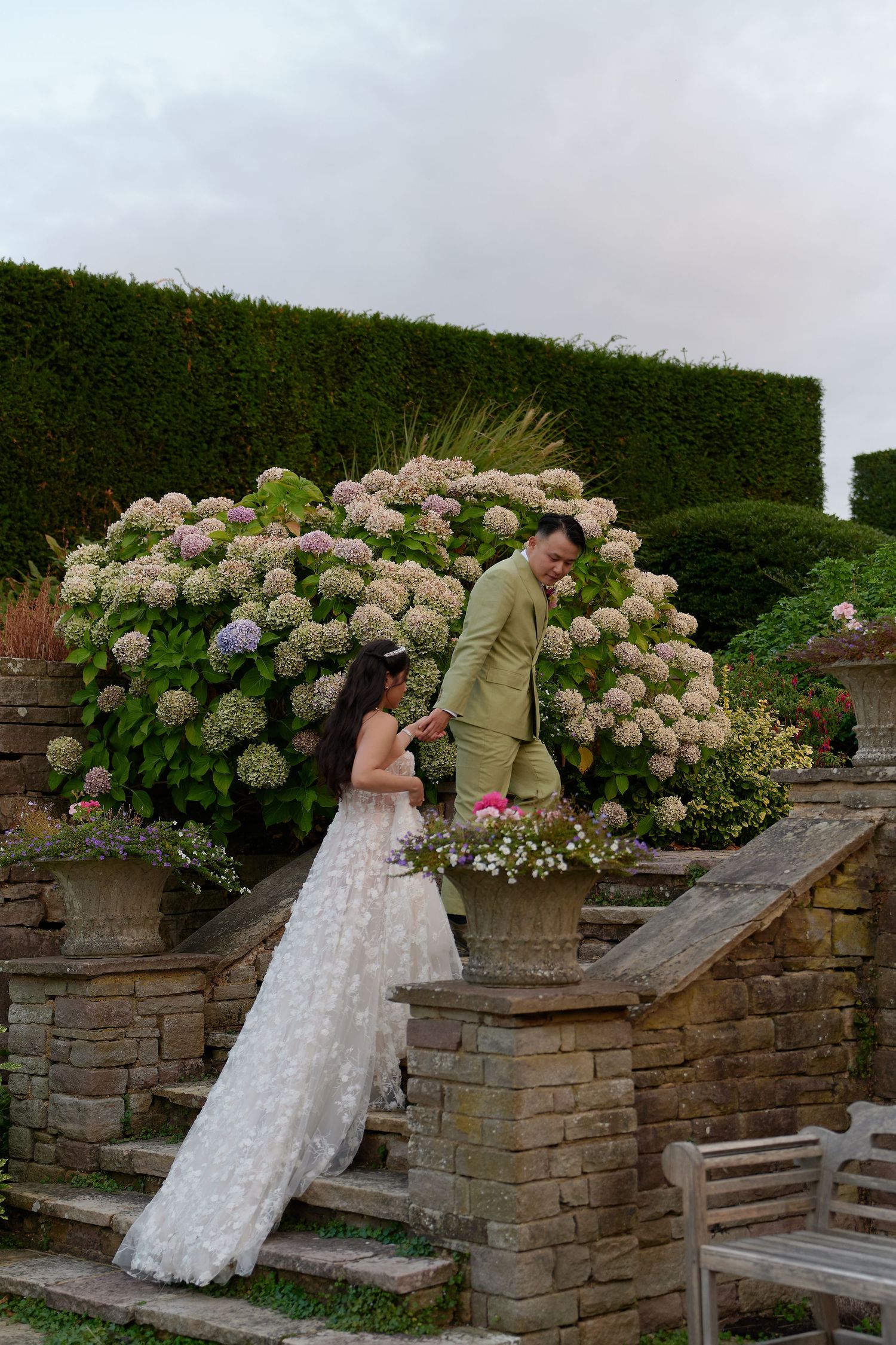 Bride in white gown and groom in sage suit walk garden stone steps surrounded by blooming hydrangeas at outdoor wedding.