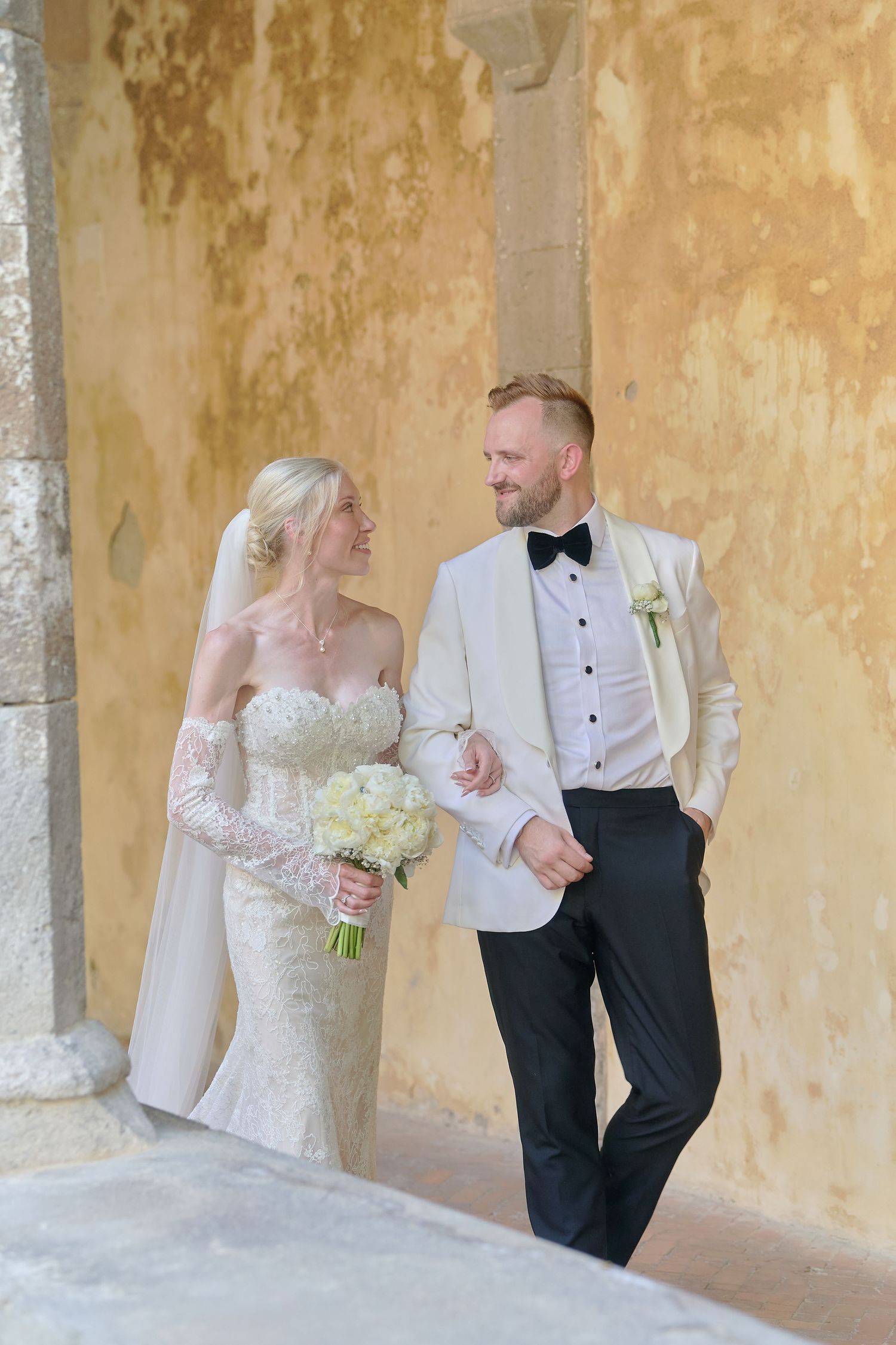 Bride and groom walking together in elegant wedding attire, she holds white bouquet, beside a rustic stone wall.