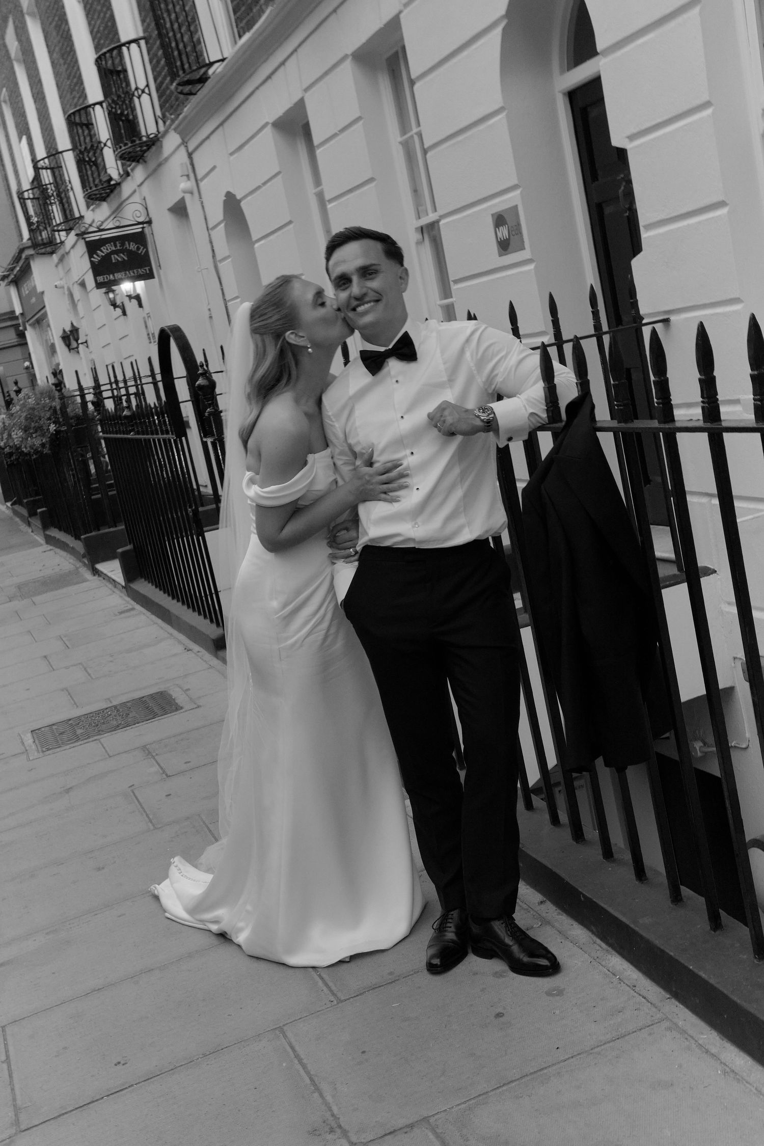 Black and white photo of a bride and groom posing on a city street, she in a white gown, he in a tuxedo.