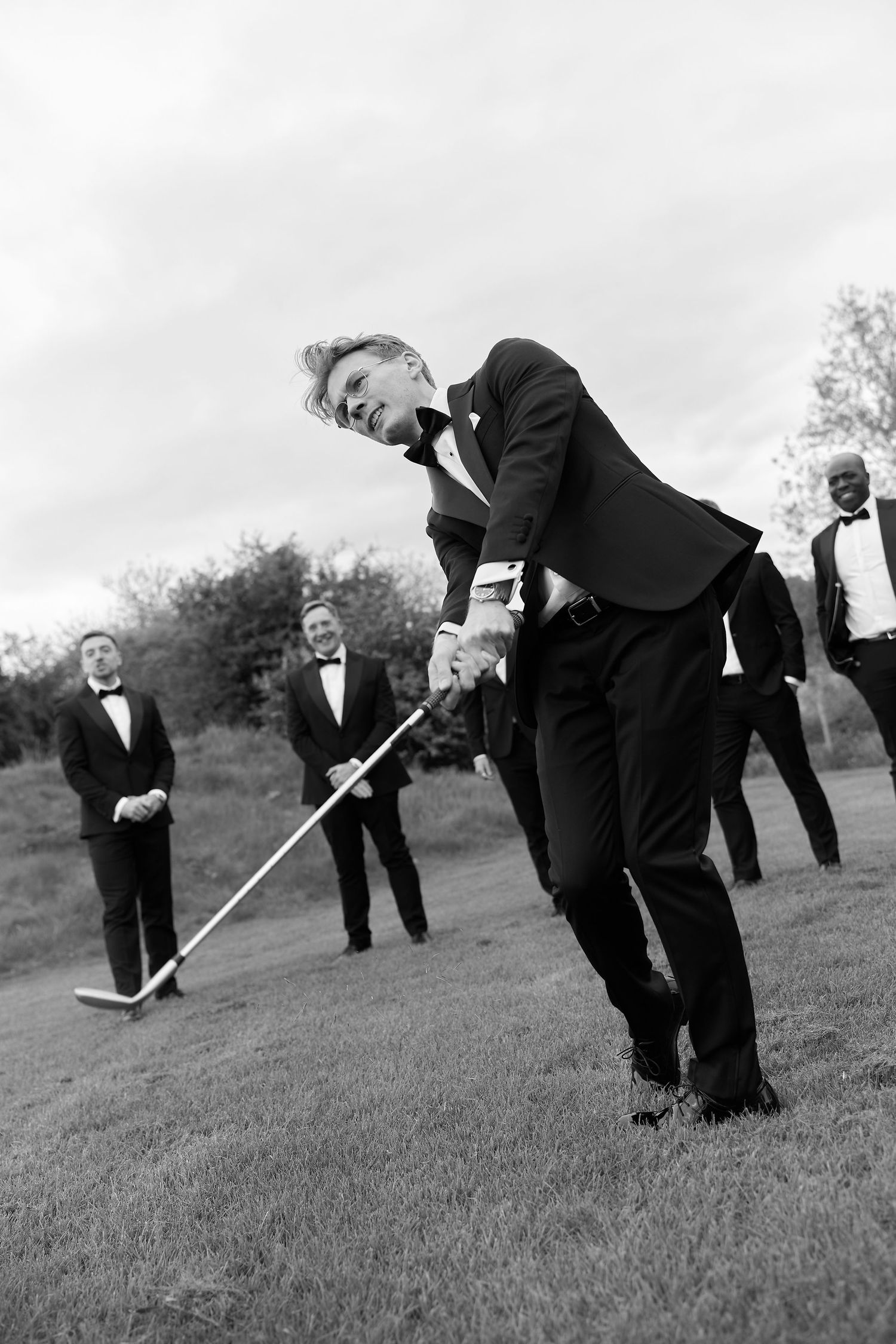 Groom swings golf club in tuxedo while groomsmen watch outdoors in black and white wedding photo.