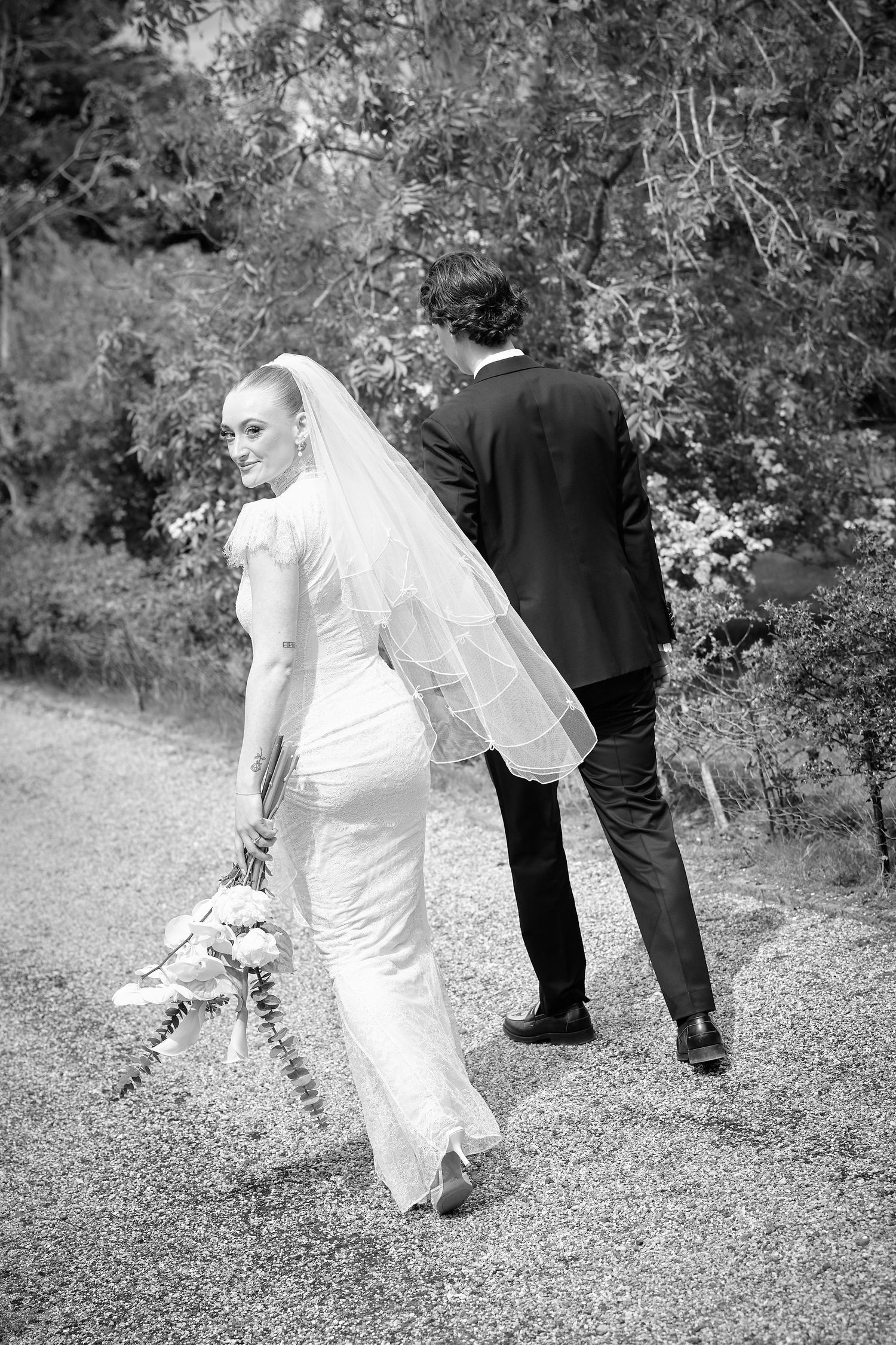 Black and white photo of a bride and groom walking on a gravel path, bride glancing back holding a bouquet.