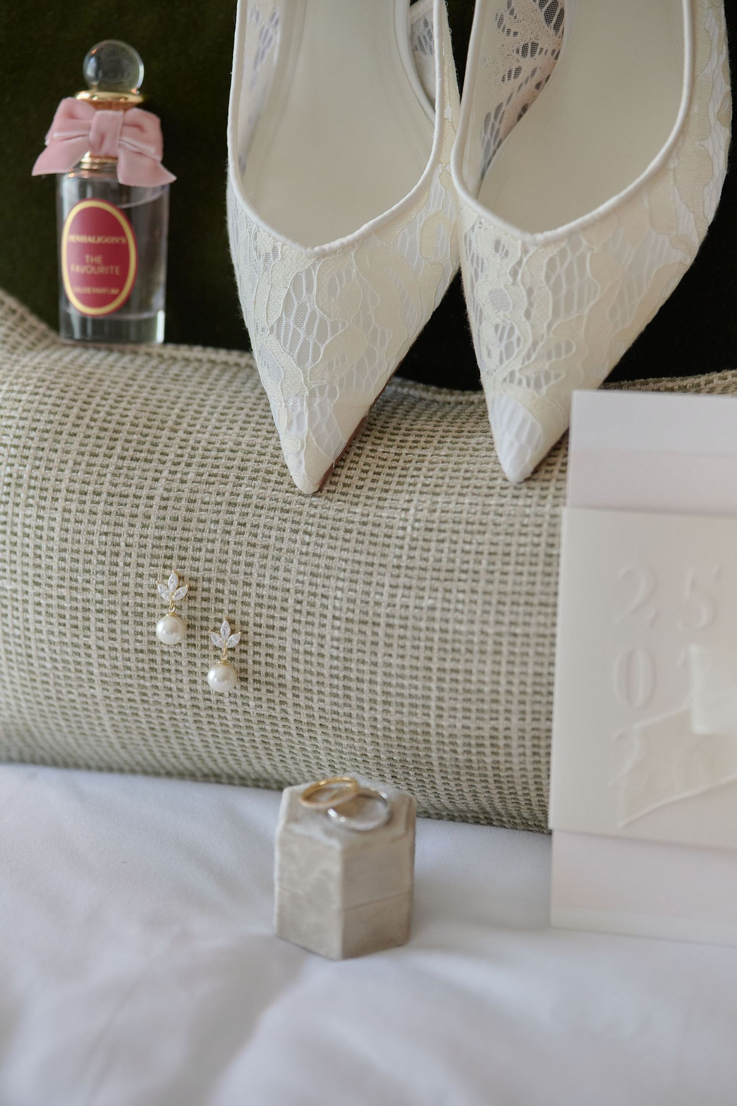 White lace bridal heels, pearl earrings, gold wedding rings, and perfume bottle arranged elegantly on a white surface.