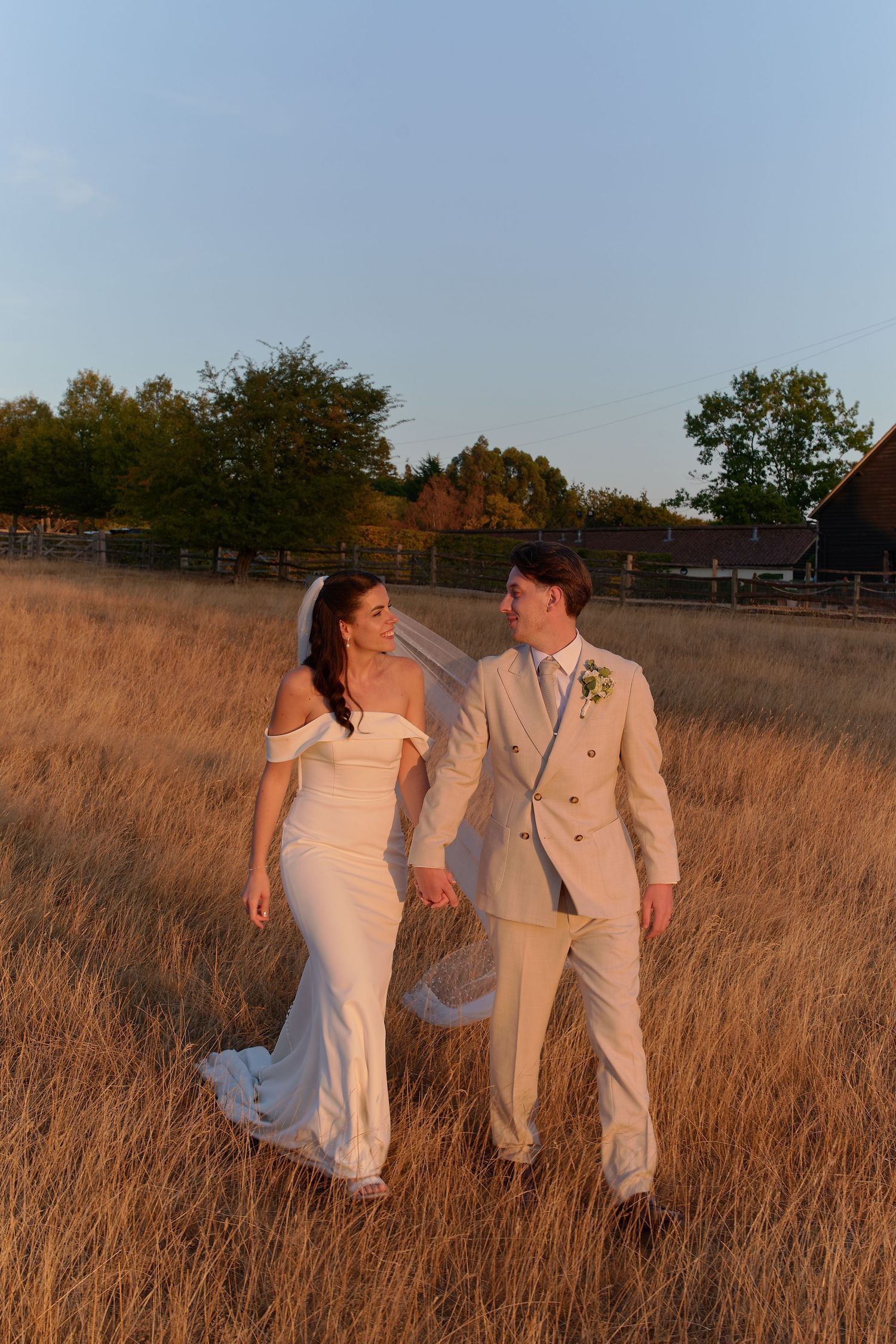Bride and groom walk hand-in-hand through golden field at sunset, she in white gown, he in beige suit.