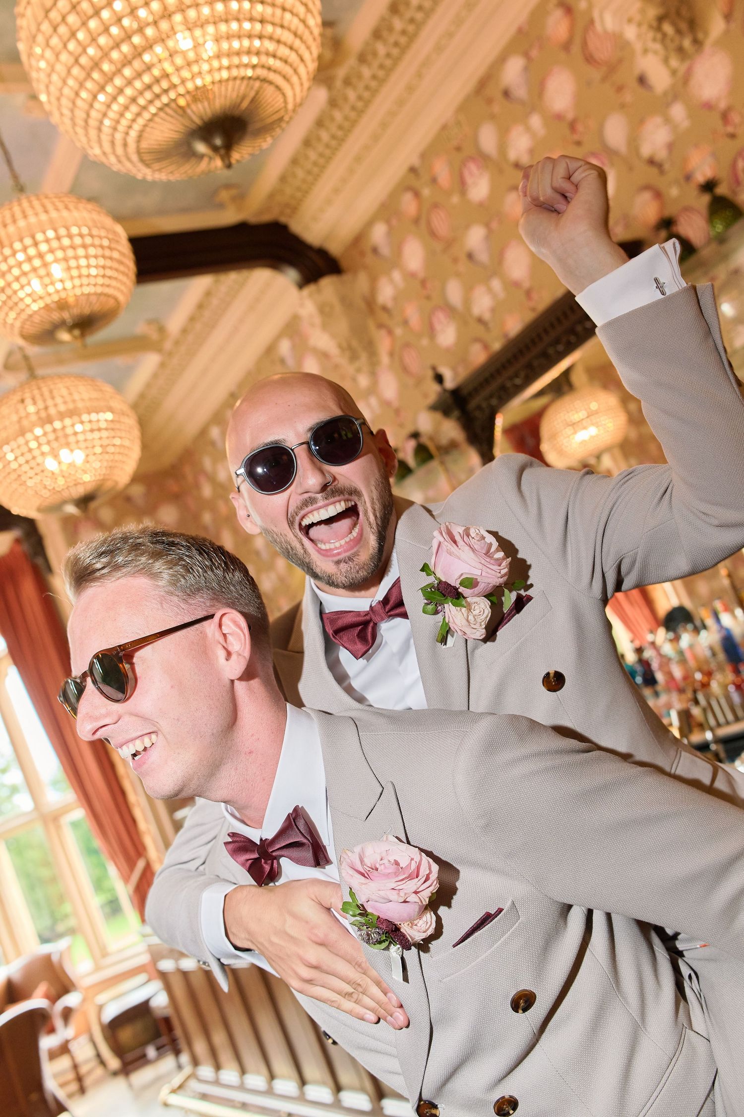 Two grooms in matching beige suits with pink boutonnieres and sunglasses celebrate joyfully at their wedding reception.