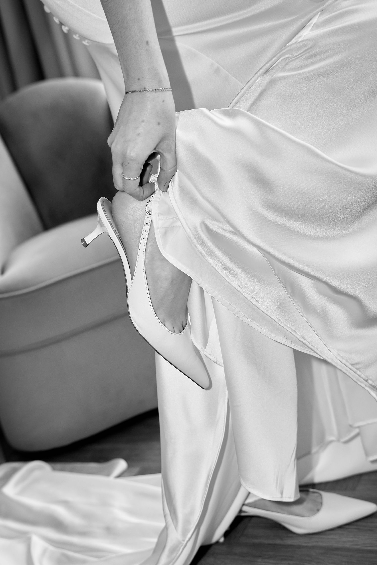 Bride in white satin gown putting on elegant white stiletto heel shoes while seated in a chair.