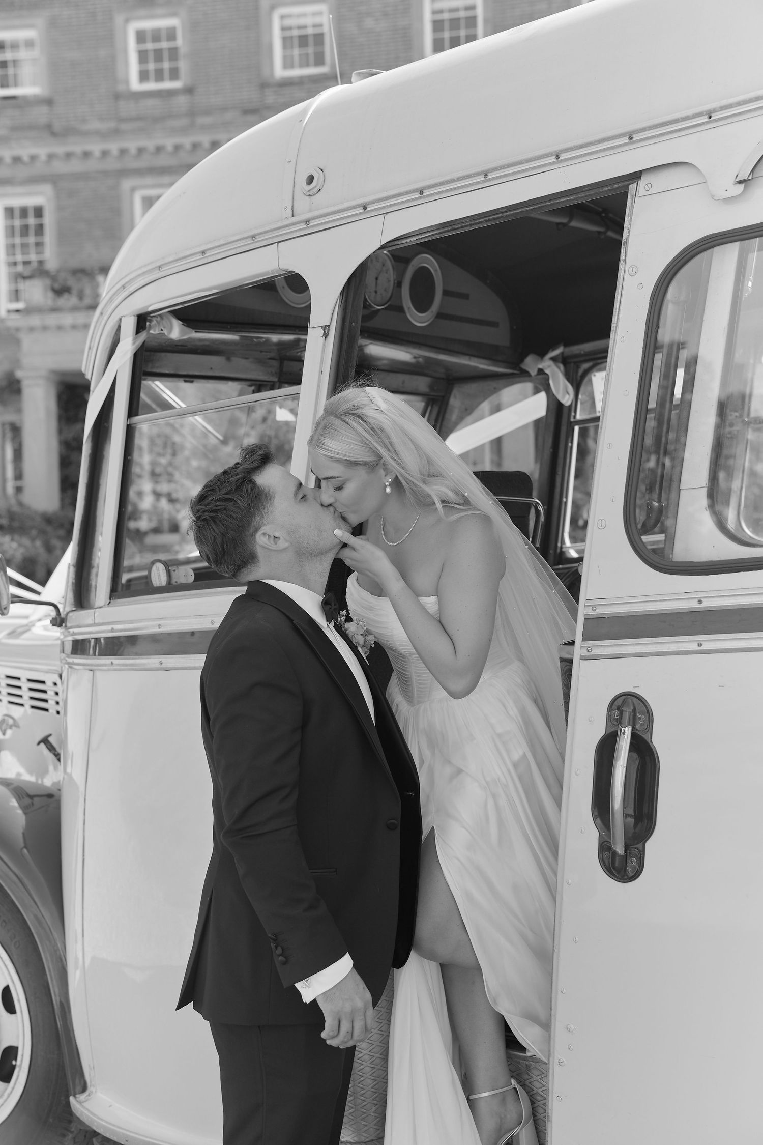 Black and white photo of bride and groom sharing a kiss beside a vintage white bus on their wedding day.