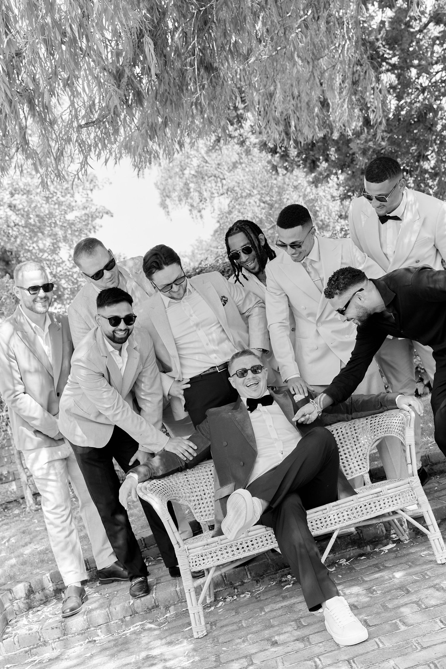 Black and white photo of a groom relaxing on a chair surrounded by smiling groomsmen in suits outdoors.