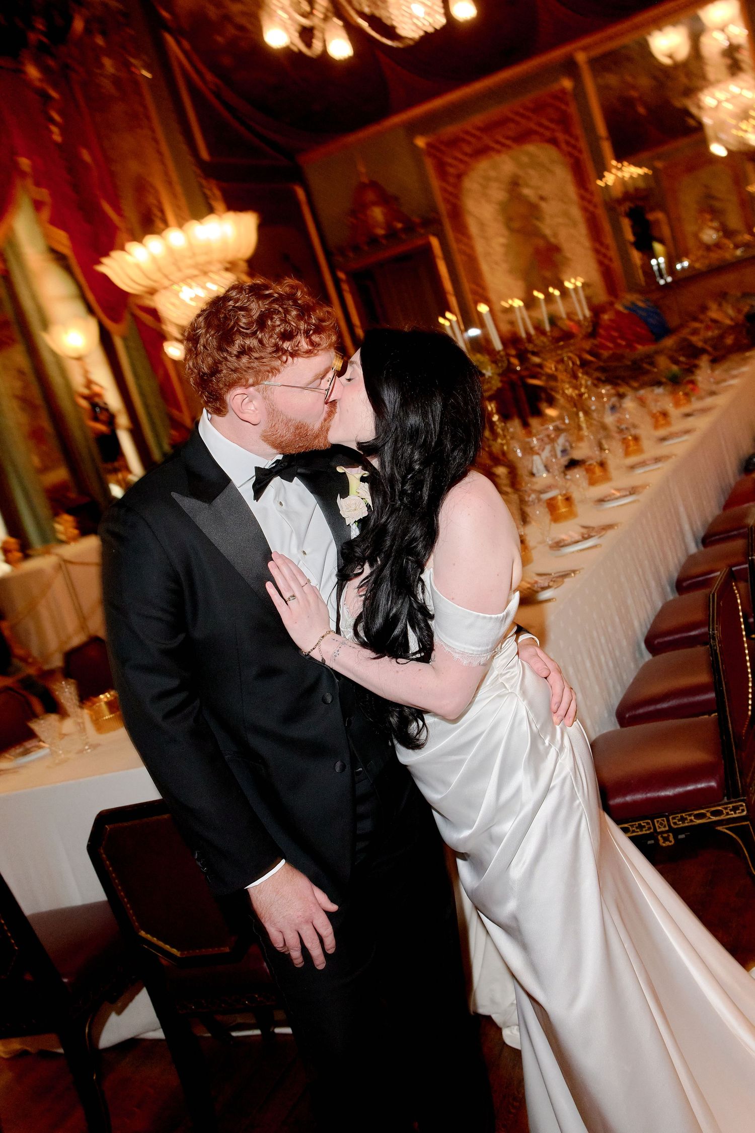 Newlywed couple sharing a romantic kiss in an elegant wedding reception hall with warm chandelier lighting.