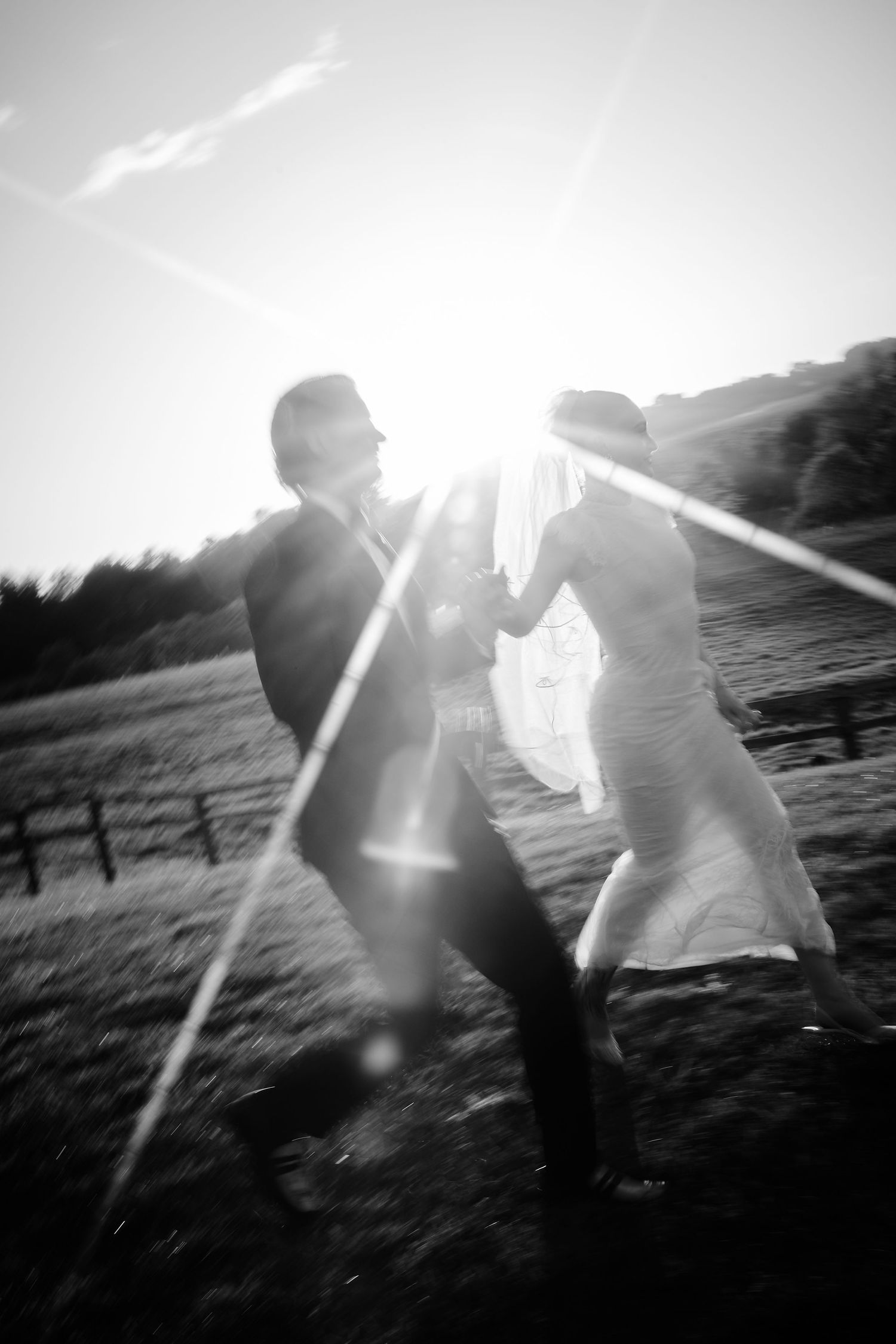 Black and white wedding photo of bride and groom dancing in sunlight with dramatic lens flare in a field.